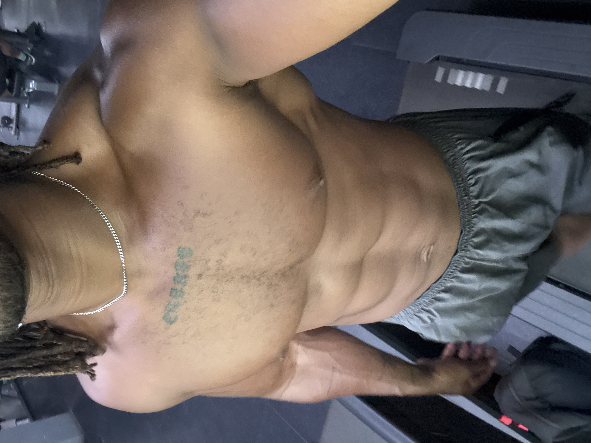 A medium shot captures a muscular, bare-chested man with dreadlocks and a silver necklace, standing on a treadmill. A tattoo is visible on his chest. He wears gray shorts and appears to be exercising. The location is Unknown, Unknown.