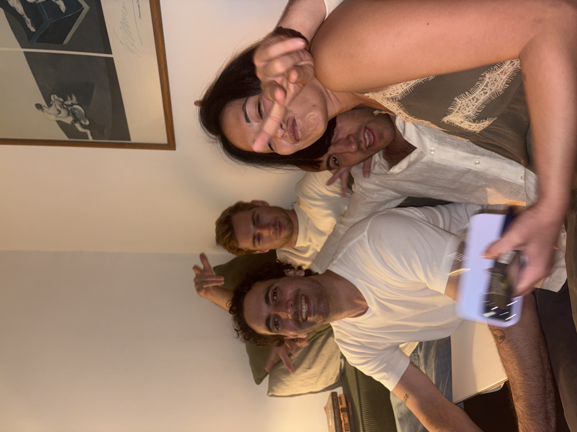 The image shows four individuals, two men and two women, in a dimly lit indoor setting, possibly a living room. The individuals are seated close together on a couch. The man in the foreground on the left has curly dark hair and is smiling. He is wearing a white t-shirt. A book is visible on the left side of the image, with its spine facing the camera. Behind him, another man with blond hair is visible. To the right, a woman is making a peace sign towards the camera, her face partially obscured. Another person's arm is wrapped around her. A painting with figures is hanging on the wall in the background. The overall impression is a casual, social gathering. No specific landmarks or indicators place this scene in Tel-Aviv, Israel.