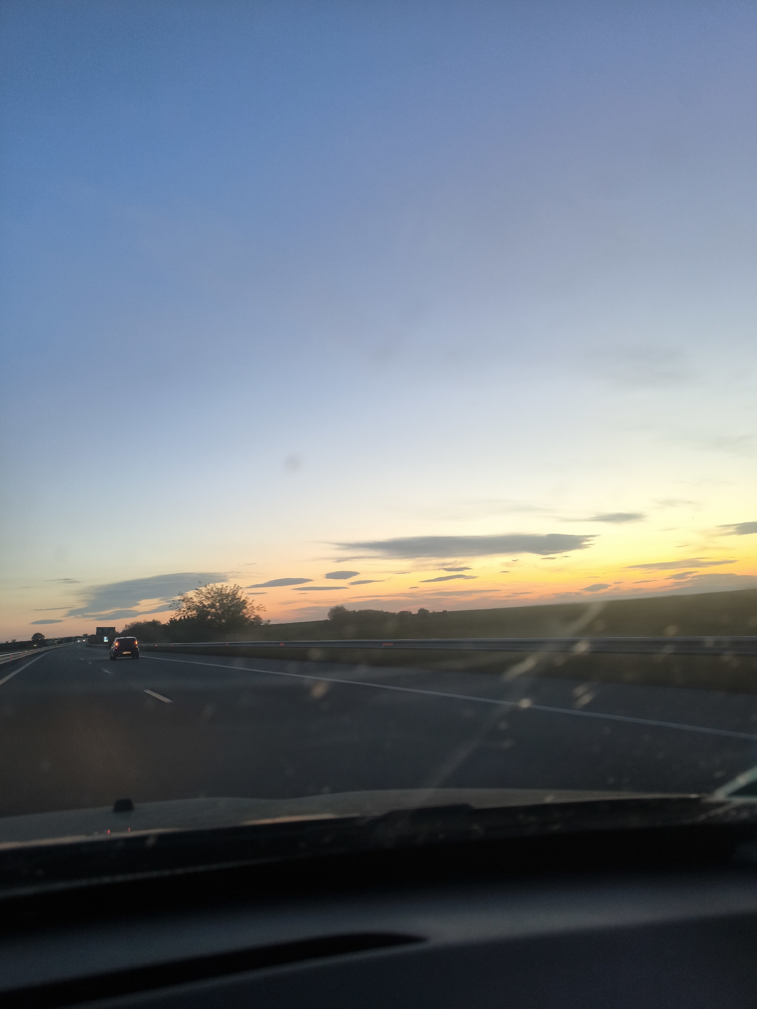 The image shows a view from inside a car driving on a highway at dusk. The sky transitions from blue at the top to orange and yellow near the horizon. There are some scattered clouds. The road is a multi-lane highway with white lines marking the lanes. Other vehicles, including a car and a truck, are visible in the distance. The landscape on either side of the highway appears to be fields and trees. The dashboard and windshield of the car are visible in the foreground. It is impossible to determine from this image if the location is in the city of Cherna Gora, Bulgaria.