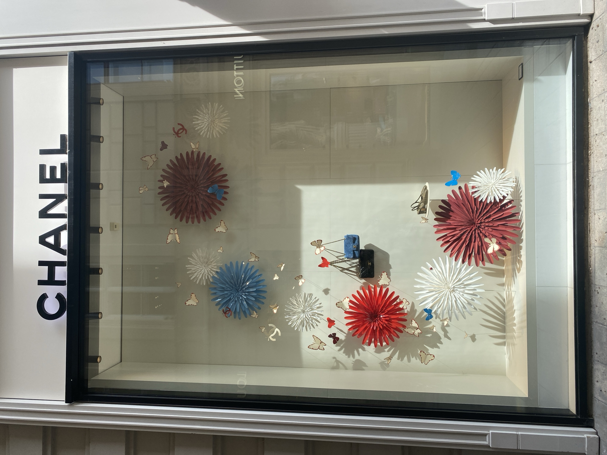 A Chanel store window display is shown in Vienna, Austria. The display features a central theme of red, white, and blue paper flowers, resembling chrysanthemums, interspersed with numerous white paper butterflies and a few blue butterflies. Two Chanel handbags, one black and one blue, are suspended in the upper right section of the display. A pair of gold heels are also visible on a small white shelf in the upper right. The Chanel logo is subtly incorporated as a metallic embellishment on some of the paper flowers. The background of the display is a light beige, and reflections of the surroundings, including architectural elements and another store with "NOTTIU" visible, are present on the glass. The exterior of the store features a white facade with vertical fluted architectural details.