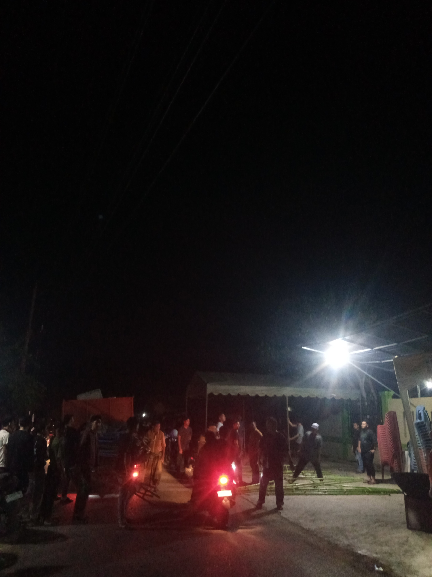The image depicts a nighttime scene in Paya Linteung, Indonesia, with a gathering of approximately 15-20 individuals. A bright spotlight illuminates a portion of the scene, highlighting several people standing and interacting. Some individuals appear to be engaged in setting up or dismantling an event space, with elements of a tent structure and stacked chairs visible. A motorcycle with its brake lights illuminated is present in the foreground. The environment appears to be a road or open area, with the darkness of the night sky above, dotted with faint power lines.