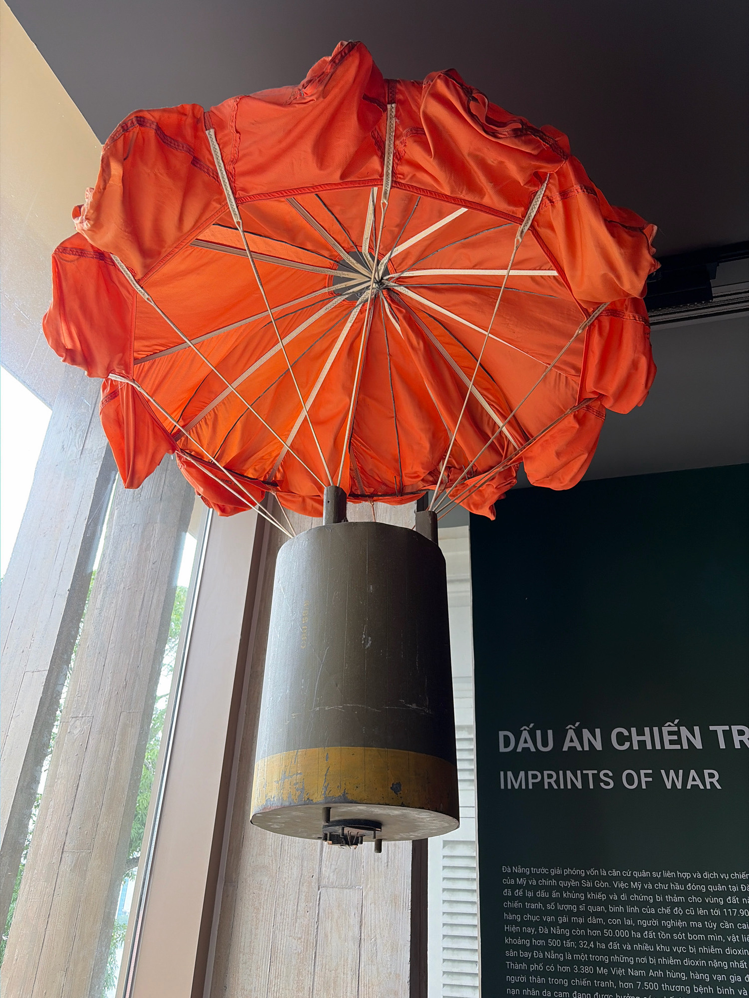 The image shows a large, open, orange parachute suspended in mid-air. Attached to the center of the parachute is a cylindrical object, which appears to be a bomb or missile casing. The casing is dark olive green with a yellow band near the bottom. The parachute and casing are displayed indoors, likely as an exhibit. In the background, a dark green banner with white text is visible. The text on the banner reads "DẤU ẤN CHIẾN TR" and "IMPRINTS OF WAR". The setting appears to be a museum or exhibition space, possibly related to the Vietnam War. To the left of the parachute, a large window reveals a blurred background of trees and what looks like a building with concrete pillars.