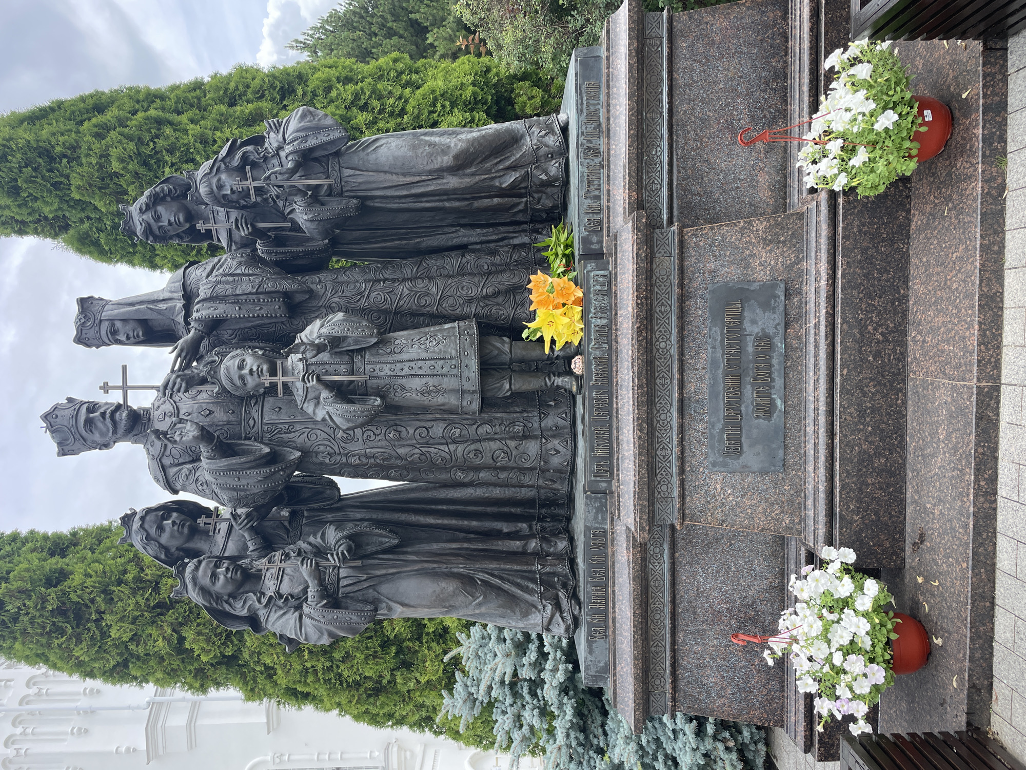 The image depicts a bronze sculpture of a family group standing on a granite pedestal in front of cypress trees and a white building, likely a church. The sculpture features a male and female figure at the center, presumably the parents, with four children arranged around them. All figures are adorned with crowns and hold crosses. The male figure wears a crown and a long, ornate robe. The female figure also wears a crown and a similar robe. The children, two boys and two girls, are dressed in historical attire and also hold crosses. A bouquet of orange and yellow flowers rests at the base of the sculpture. Two potted plants filled with white flowers are placed on the lower level of the pedestal. Inscriptions are visible on the pedestal, indicating names or historical references. The environment suggests an outdoor setting, likely a memorial site. The overall scene conveys a solemn and historical atmosphere, suggesting a monument dedicated to a significant family. The city of Diveyevo, Russia, is known for its religious sites, including the Diveyevo Monastery, which often features historical and religious monuments.