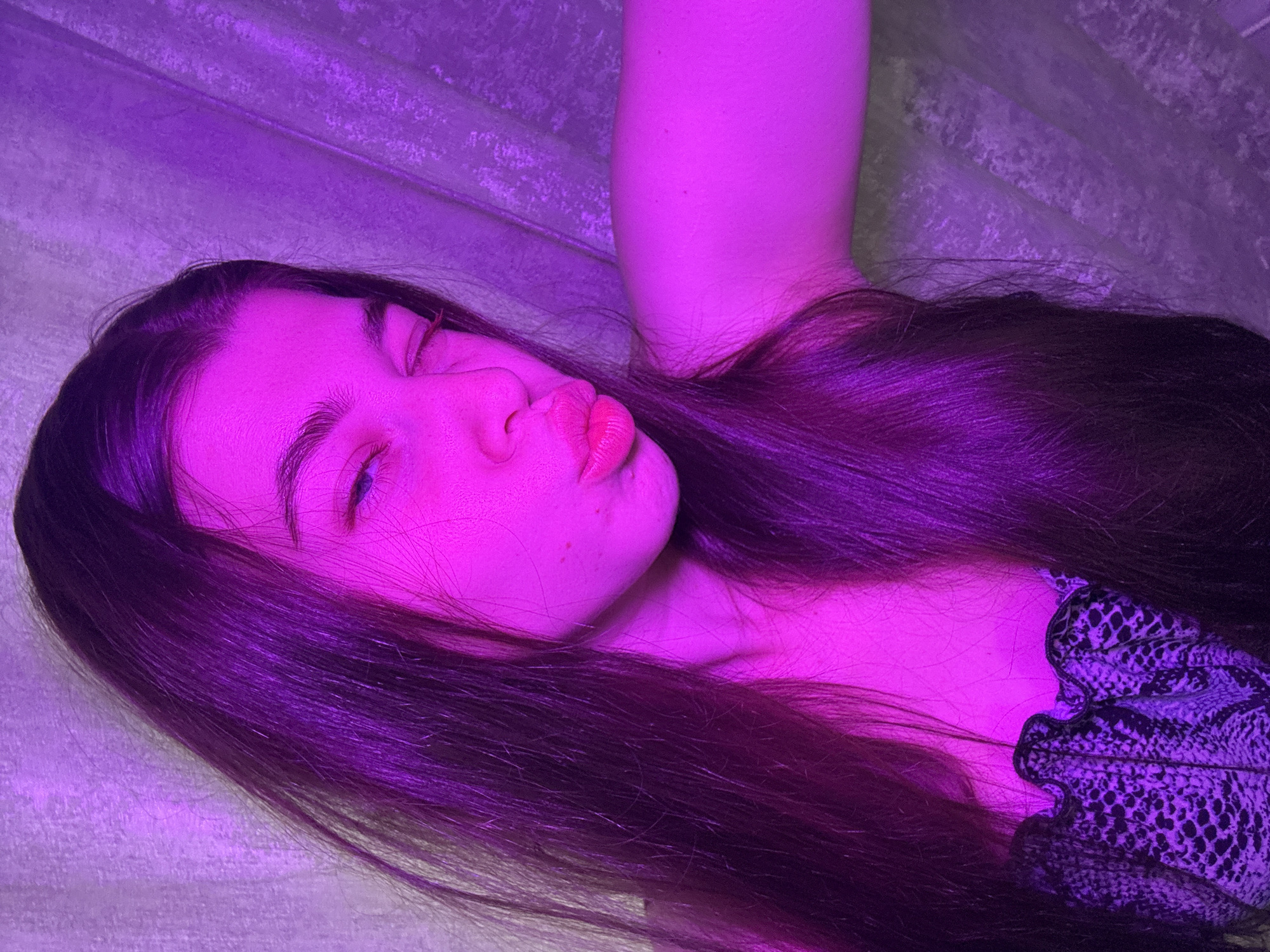 A young woman with long, dark hair is lying down. She is pouting with her lips puckered. Her hair is spread out around her. She is wearing a patterned top with lace detailing. The scene is illuminated with a strong purple light. The background appears to be a wall with a textured surface. There is no information about the location of the subject being in Мещёры, Russia.