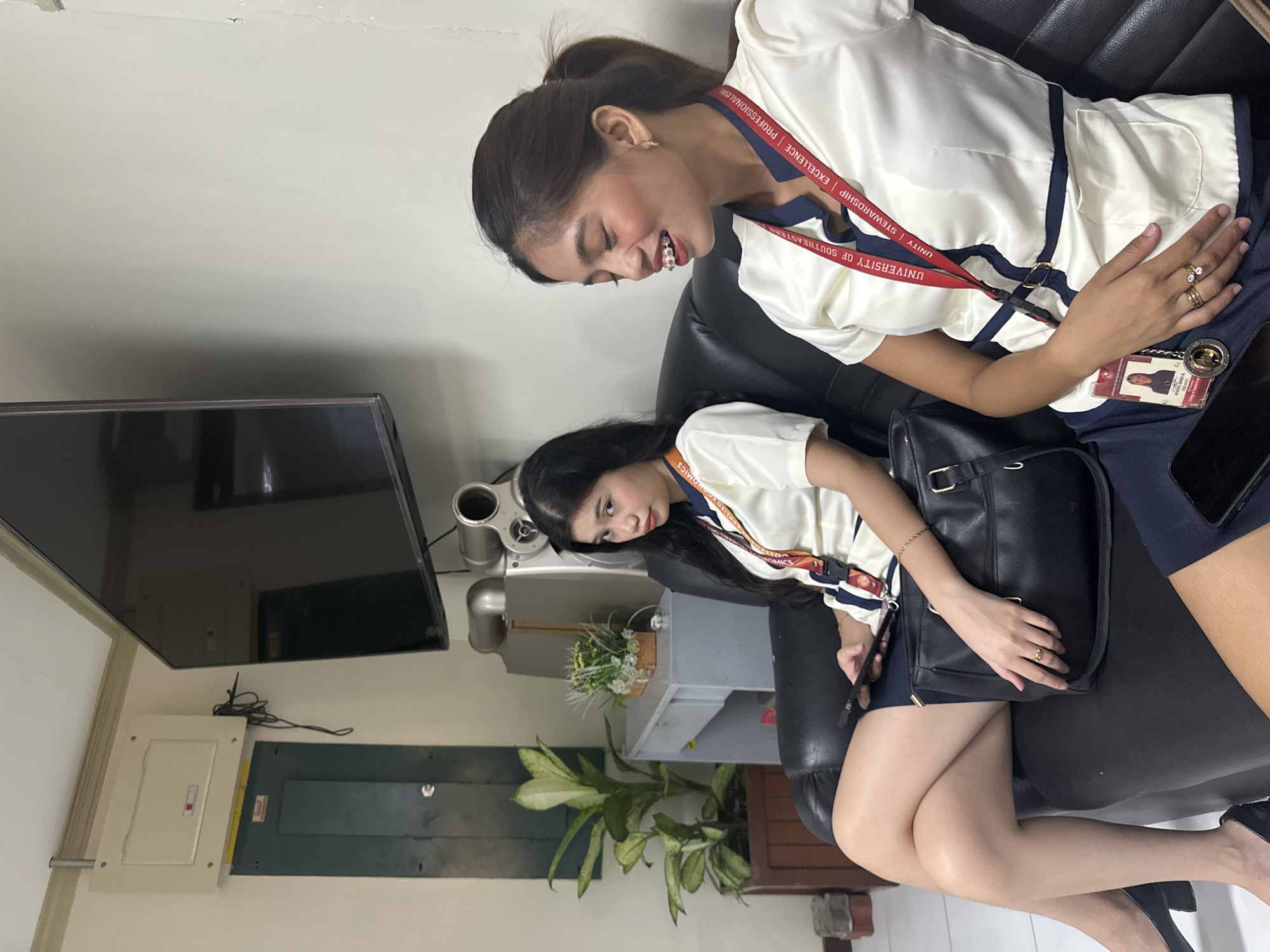 Two young women are seated on a black leather couch, indoors. The woman on the left is looking towards the viewer with a neutral expression, holding a black smartphone in her right hand. She is wearing a white blouse with navy blue trim and a navy blue skirt, along with black high heels. A black purse rests on her lap. The woman on the right is looking to her right with a wide smile, revealing braces on her teeth. She is wearing a similar white blouse with navy blue trim and a navy blue skirt. Both women are wearing lanyards around their necks, which appear to be university identification badges.
The background includes a wall with a large flat-screen television mounted on it. To the left of the television, there is an electrical panel and a large green metal cabinet. In the far left of the image, a large potted plant with green and white leaves is visible. Further back, a grey desk or cabinet can be seen. The lighting suggests an indoor setting, possibly a waiting area or lounge. The presence of university-related attire implies an academic or institutional environment. The specific location in Davao City, Philippines, is not determinable from the image alone.