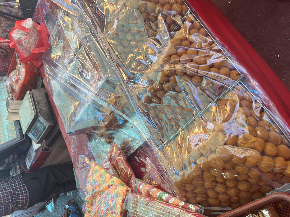 The image shows a street vendor selling sweets in Panchkula, India. The sweets are arranged in a grid pattern on a red table covered with plastic sheeting. The sweets are small, round, and appear to be a type of Indian sweet called "ladoo." There is a red plastic bag containing what appears to be dry fruits in the upper right corner of the image. There are also some other unidentified items in the lower left corner of the image. The vendor is not visible in the image.  The image is taken at a street stall in Panchkula, India, at the coordinates 30.725 latitude and 76.86 longitude.