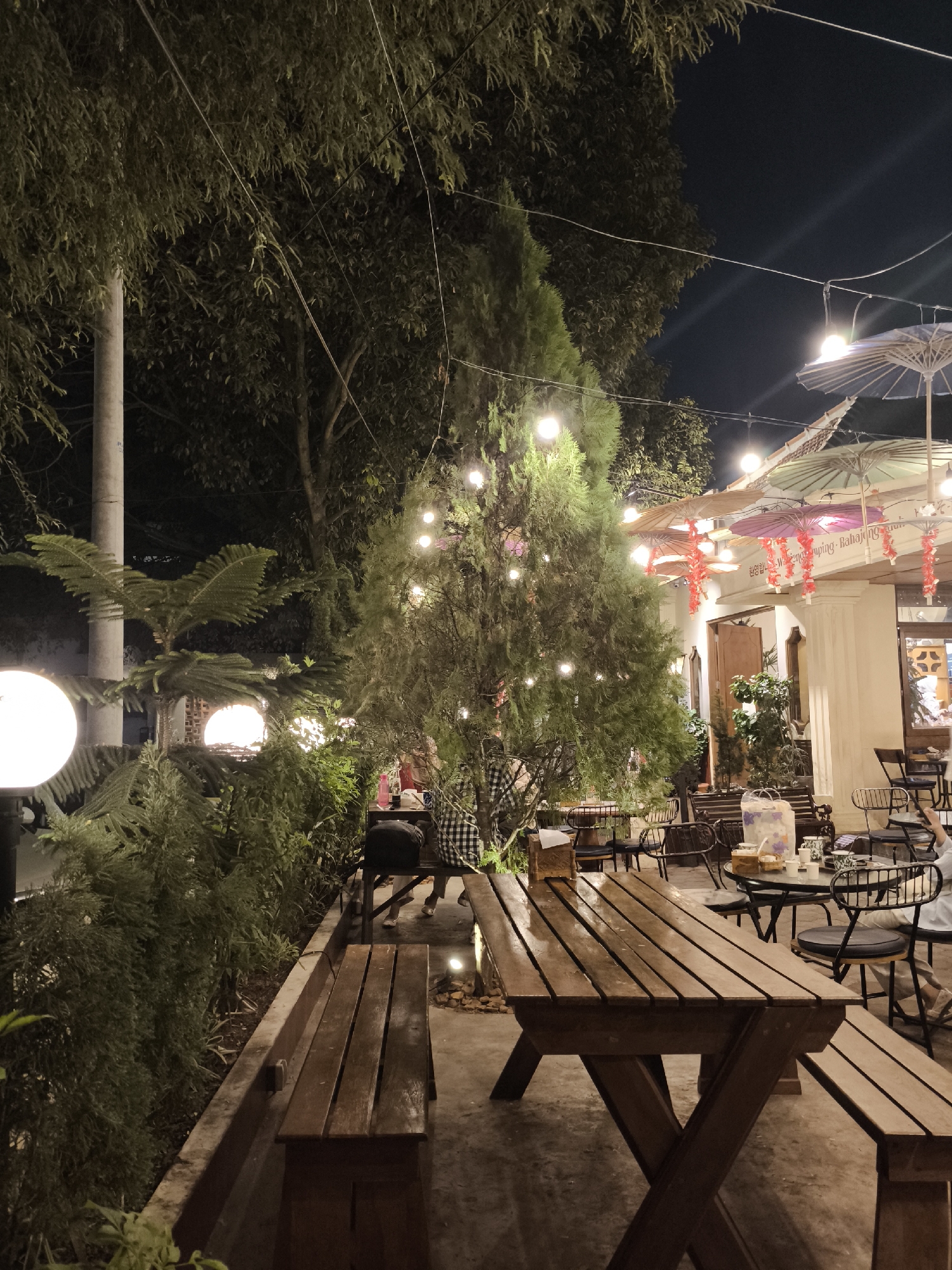 This image depicts an outdoor seating area of a cafe or restaurant at night in Surakarta, Indonesia. The scene is illuminated by string lights and large, round outdoor lamps. A prominent wooden picnic table with benches is in the foreground, appearing wet, suggesting recent rain. In the background, there is a building with an awning decorated with colorful, traditional-looking umbrellas and hanging red ornaments. Several metal chairs and small tables are arranged in front of the building. Various trees and dense greenery surround the seating area. A person is partially visible in the background, sitting at one of the tables. The overall atmosphere suggests a relaxed evening setting.