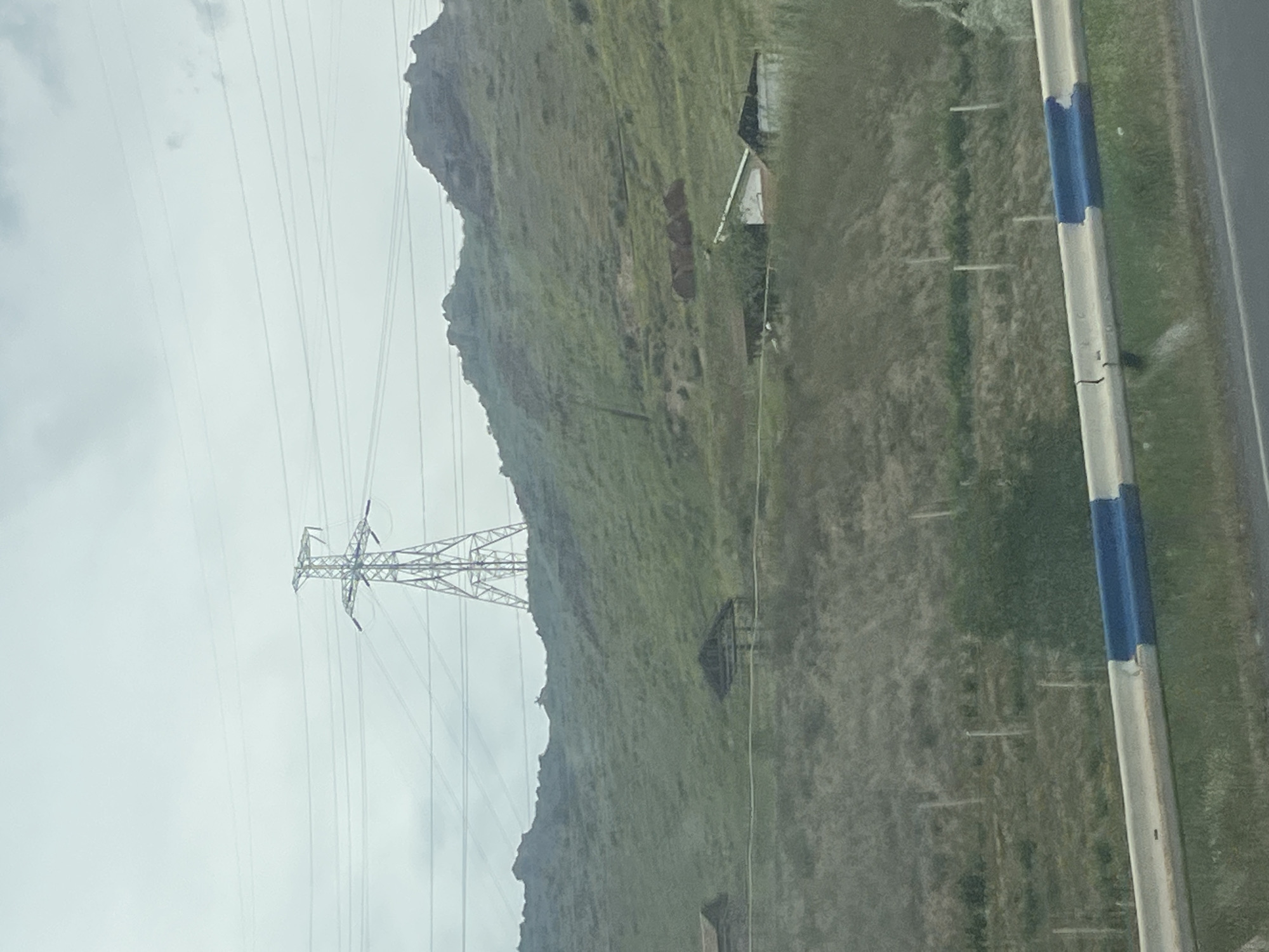 This landscape photograph captures a vast, rugged scene featuring natural terrain and human infrastructure, likely taken from a moving vehicle.
The dominant feature is a large, rolling hill or small mountain, covered primarily in green and brown grasses and low-lying vegetation. Its jagged ridgeline stretches across the middle of the frame from left to right. The sky above is uniformly light grey and overcast, indicating a cloudy day.
A prominent, tall metal electrical transmission tower (pylon) stands on the lower slope of the hill towards the left, with multiple power lines extending horizontally across the sky. At various points on the lower slopes and base of the hill, several small, simple structures, possibly shacks or outbuildings, are visible; one notably has a white roof.
On the far right, a grey road barrier or guardrail with distinct blue and white painted sections runs vertically, suggesting the vantage point is from a road. A narrow strip of asphalt road is partially visible immediately to the right of the barrier, and a grassy verge separates the road from the hill. Several faint, light-colored vertical markers are dotted in the grassy area between the road and the hill.
No discernible human or animal activity is occurring, making the scene appear static and somewhat remote. The time of day is daylight, but the absence of shadows points to an overcast sky. The context provided indicates the location is Chiva, Armenia, which aligns with the mountainous and pastoral landscape. There is no visible text in the image.