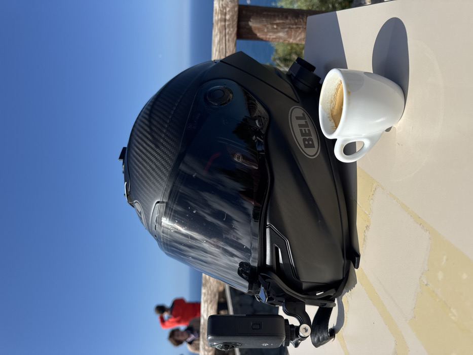 A matte black Bell motorcycle helmet, featuring a carbon fiber pattern on its crown and a dark tinted visor, rests on a light-colored, possibly tiled, outdoor table. An Insta360 action camera is mounted on the helmet's left side via a strap. A small white ceramic cup containing espresso is situated to the right of the helmet on the same table. The scene is set at a coastal viewpoint in Deià, Spain, overlooking the deep blue Mediterranean Sea under a clear blue sky. A rustic wooden railing runs horizontally across the mid-ground. Two blurred individuals are visible in the distant background behind the railing; one wears a red top and appears to be observing the sea.