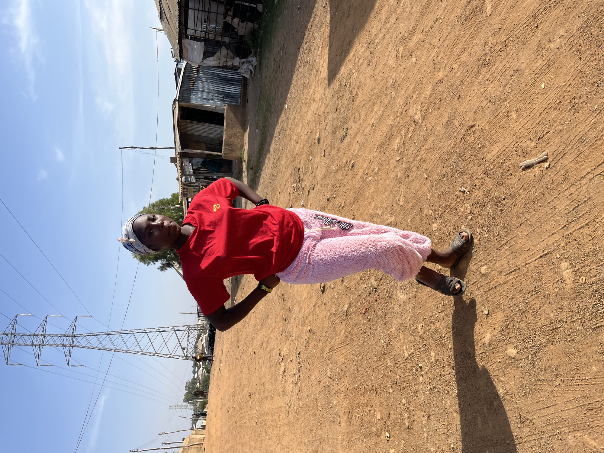 A young woman stands on a dirt road in Jos, Nigeria. She is wearing a red T-shirt, fuzzy pink pants, and sandals. A blue and white headscarf covers her hair. She has her hands on her hips. Behind her are dilapidated buildings and an electrical tower. The sky is blue with some clouds. The ground is dry and brown.