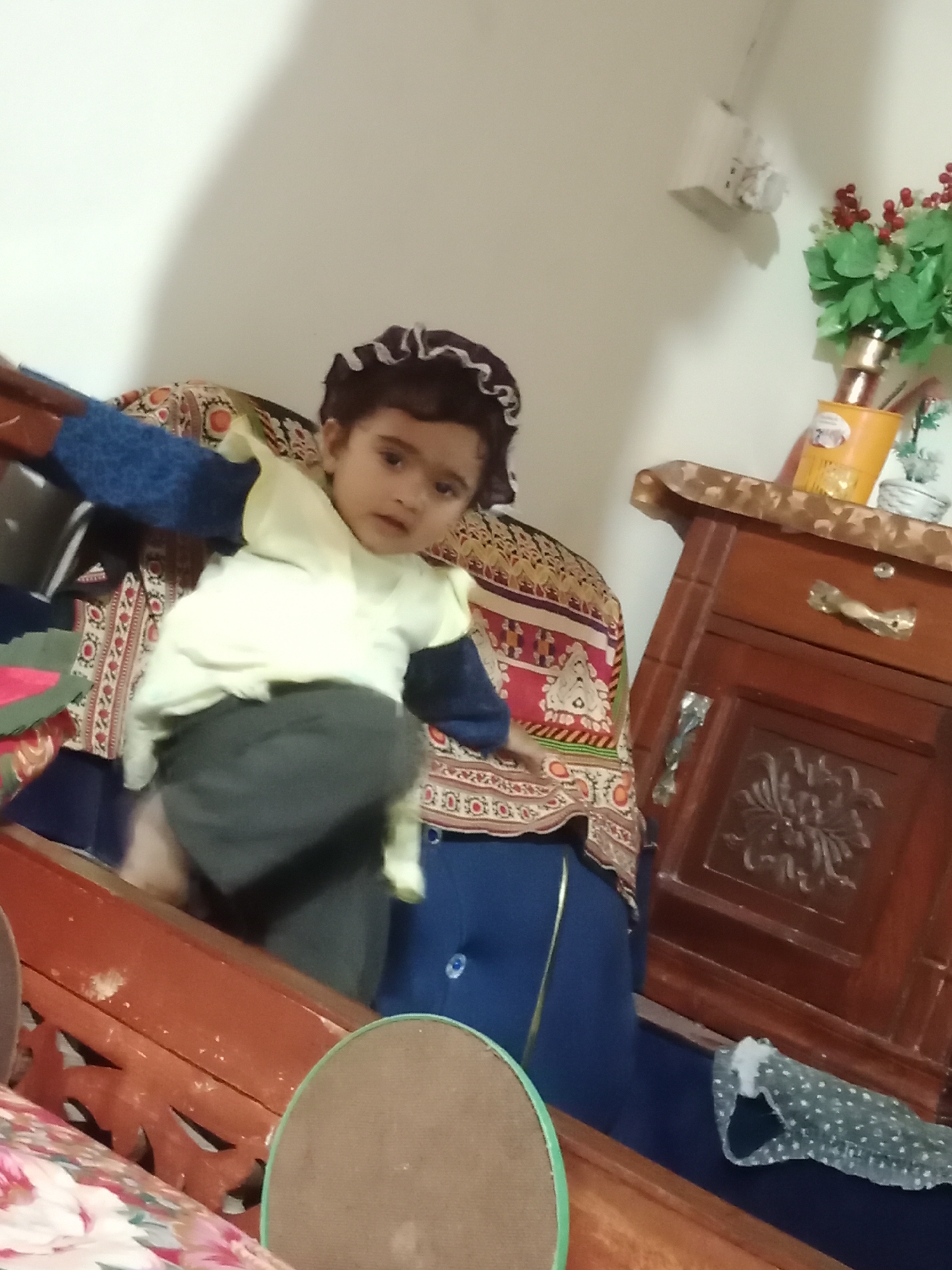 A child in a ruffled cap stands on a floral patterned cushion on what appears to be a piece of wooden furniture. The child wears a light-colored top over a blue-sleeved shirt and dark pants. Behind the cushion is a wall painted in a light color. To the right is a wooden cabinet with a drawer and a decorative floral design. Atop the cabinet are a plant and a yellow container. The photograph seems to be taken indoors, possibly in a residence. The specific location within the city of Karamsar, Pakistan is indeterminable from the image.