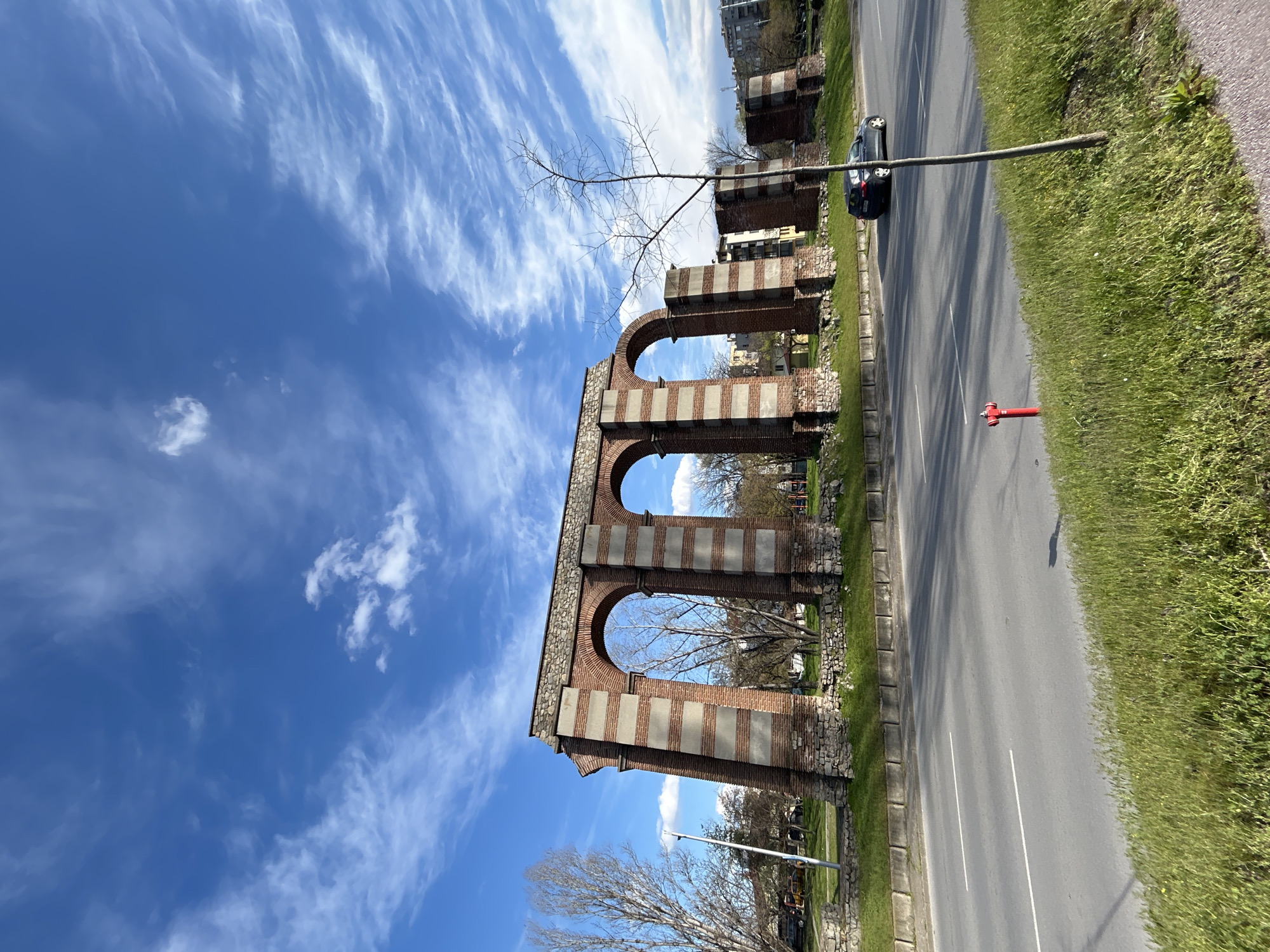 The image shows the remains of an ancient Roman aqueduct in Plovdiv, Bulgaria. The structure consists of three large brick and concrete arches, partially intact. The aqueduct is situated alongside a road with a passing black car and a red fire hydrant. The sky above is blue with streaky white clouds. Adjacent to the road is a grassy area. Bare trees are visible near the aqueduct. There are also some buildings in the background.