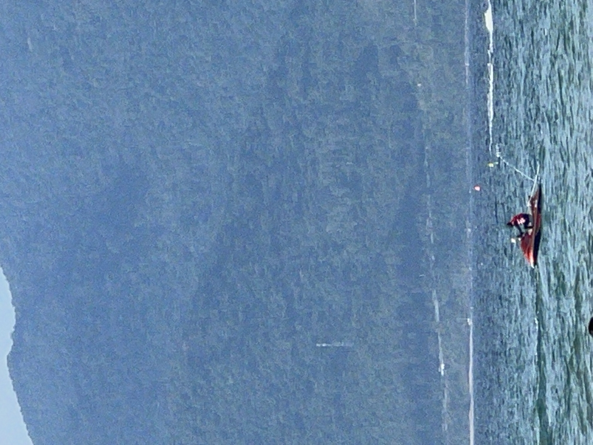 A wide shot captures a serene seascape with a mountainous backdrop, likely in Da Nang, Vietnam. The water is a deep blue, with gentle waves creating whitecaps. In the foreground, a jet ski is being ridden by two individuals. The jet ski is red and white and is cutting through the water, leaving a white wake. The two riders are wearing dark clothing. Further out on the water, a buoy is visible, marking a boundary or course. In the distance, a long, thin strip of land with some buildings and vegetation separates the water from a large, densely forested mountain range that dominates the horizon. The sky is a light, hazy blue. The overall impression is one of a peaceful and beautiful natural landscape, with recreational activity adding a touch of life.