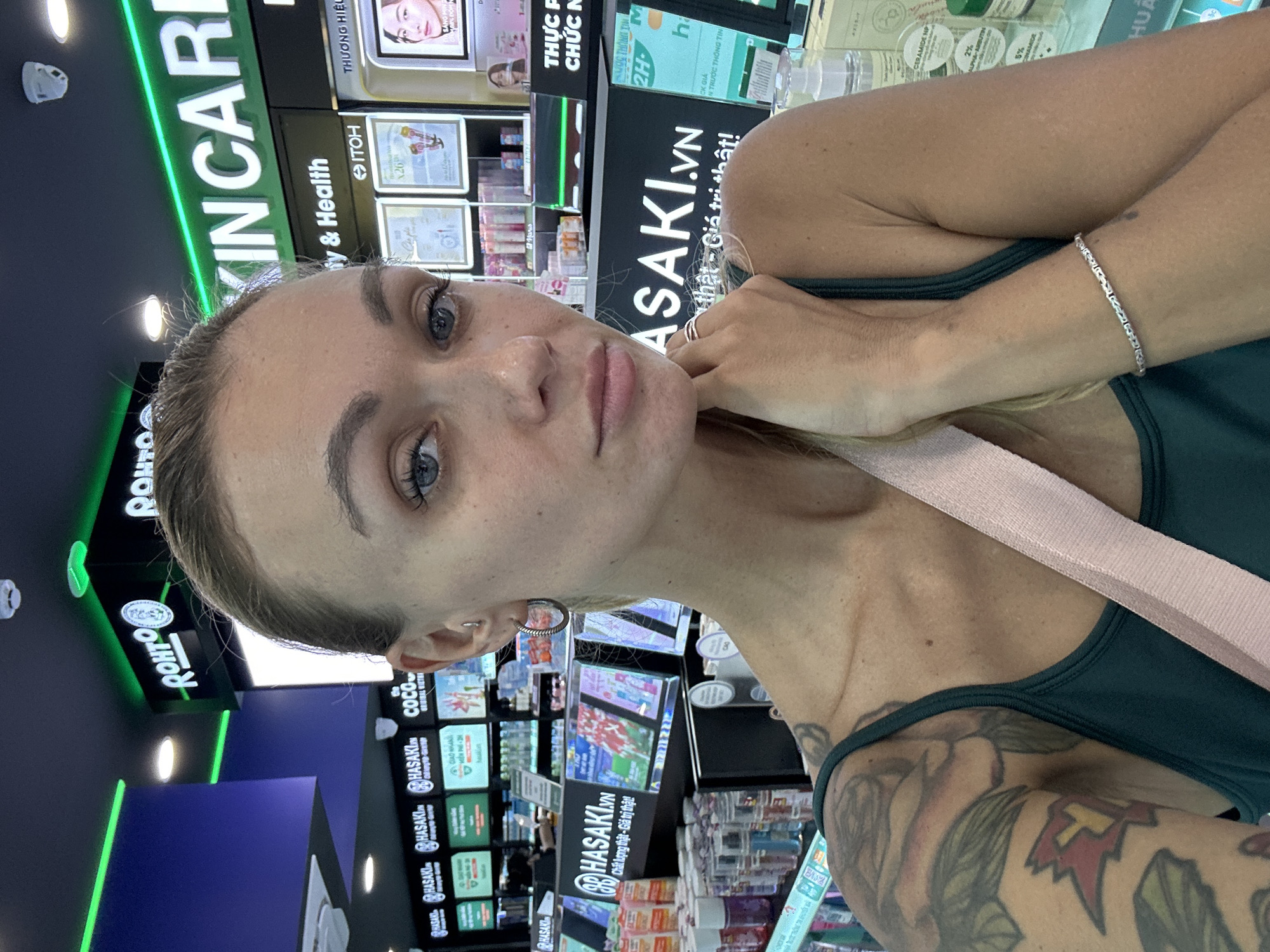 The image shows a woman posing for a selfie in a beauty store. The background reveals shelves filled with skincare products, primarily from the brand "Hasaki.VN." Vietnamese text and product labels are visible. The location is suggested to be in Phan Thiết, Vietnam. The woman is wearing a dark green tank top and has a pink strap across her chest. She has light skin, blonde hair, and a tattoo on her upper arm. She's wearing a bracelet and rings.