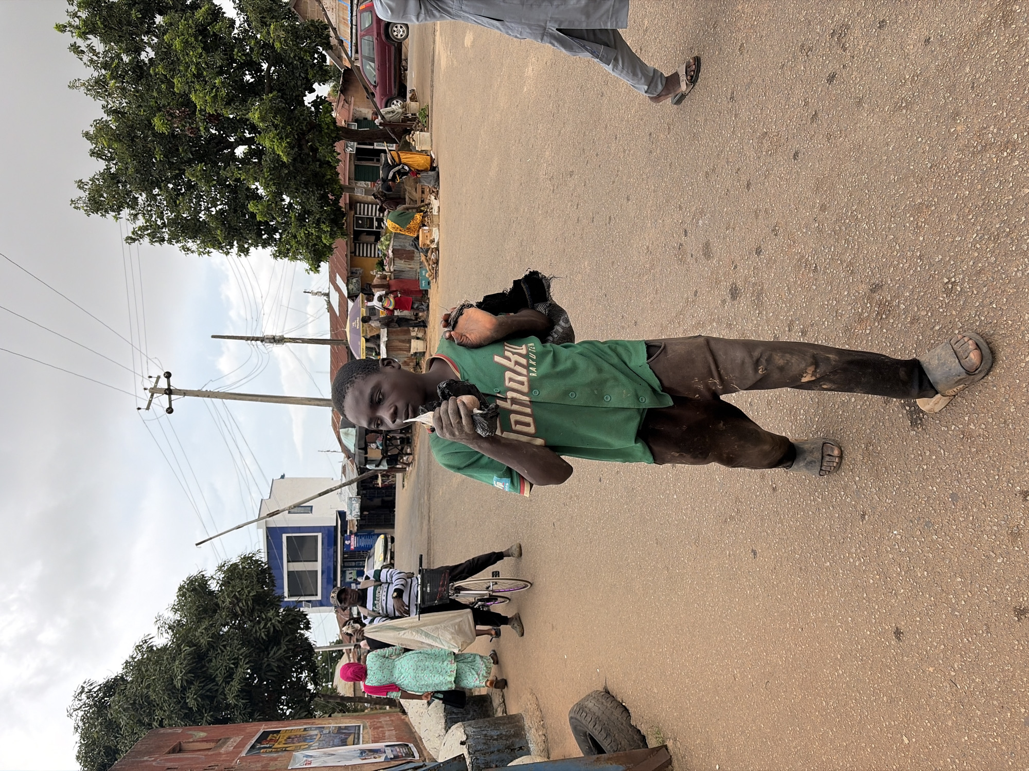 In an outdoor urban scene, likely in Jos, Nigeria, a young boy walks towards the viewer on a dusty, asphalt road. He wears a green short-sleeved baseball jersey with "JOHO" and "RAKUTEN" written on it, and dark brown, dirt-stained pants. His feet are clad in dark, worn sandals. He holds a white object, possibly food or candy, in his right hand and a crumpled black plastic bag in his left. His expression is neutral.
To the left, a woman in a light blue and white patterned dress and a pink headscarf walks away from the camera, carrying a white bag. Beside her, a person in a black and white striped shirt and a dark baseball cap stands with a bicycle, also facing away. A maroon-colored car is parked on the right side of the road in the background. Numerous market stalls with tin roofs are visible, with people tending to goods and interacting. A large, leafy tree stands on the right. The sky is overcast. The overall impression is of a busy, somewhat chaotic marketplace environment.