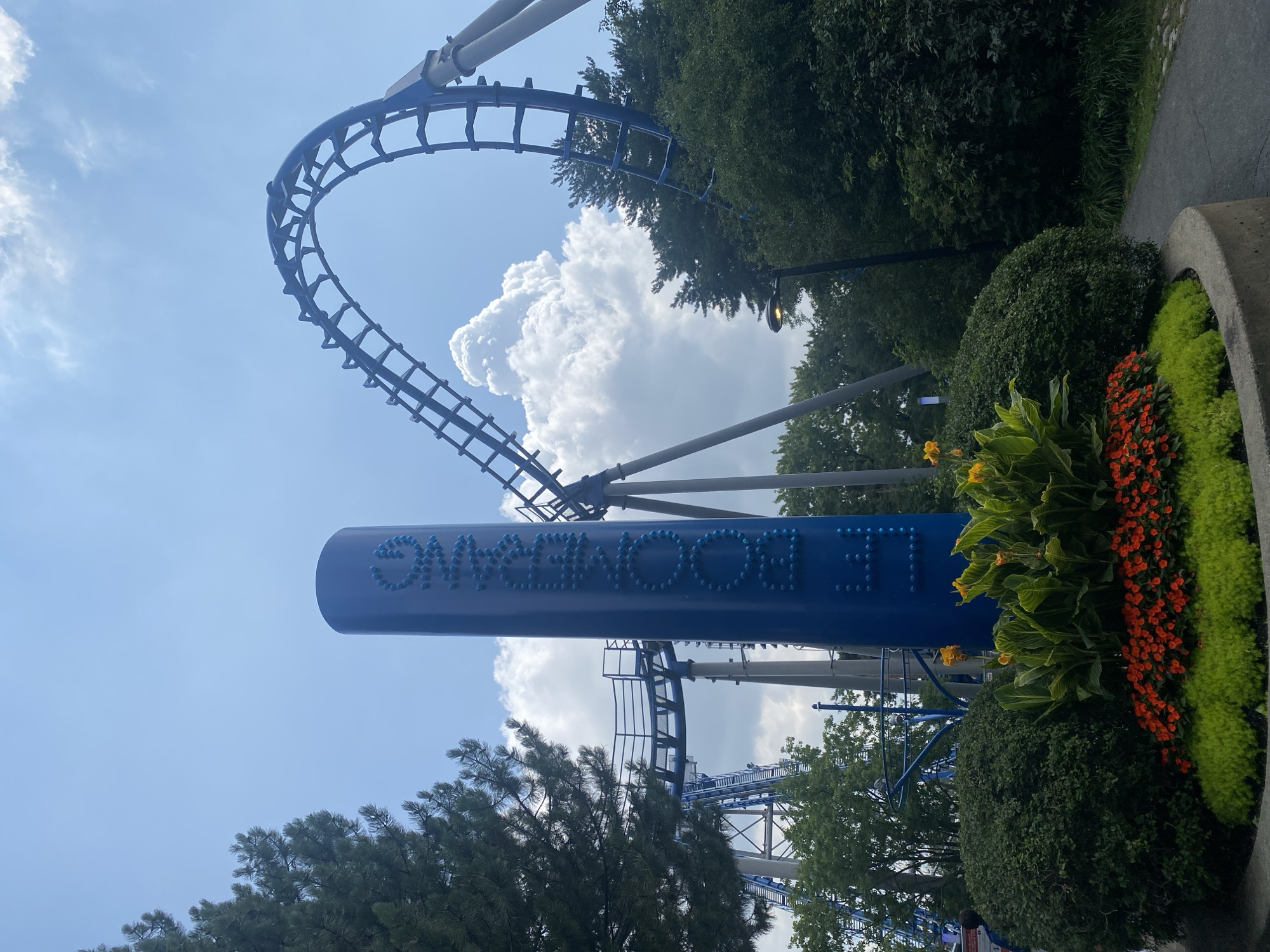 The image captures an outdoor scene featuring a blue roller coaster with a loop against a bright blue sky with scattered white clouds. In the foreground, a tall, cylindrical blue sign with the word "BOOMERANG" written vertically in raised, light blue letters stands prominently. The sign is adorned with small, light blue spheres forming the letters. To the left of the sign, a lush green evergreen tree is visible. In front of the sign and tree, there is a flower bed with a mix of vibrant red and green foliage.
The roller coaster track is elevated, supported by gray pillars, and appears to be part of an amusement park. The overall environment suggests a sunny day in an outdoor recreational area. The image's aesthetic points to an amusement park in Montreal, Canada, given the prominence of the "Boomerang" roller coaster, a known attraction at La Ronde, a Six Flags amusement park located on Île Sainte-Hélène in Montreal.