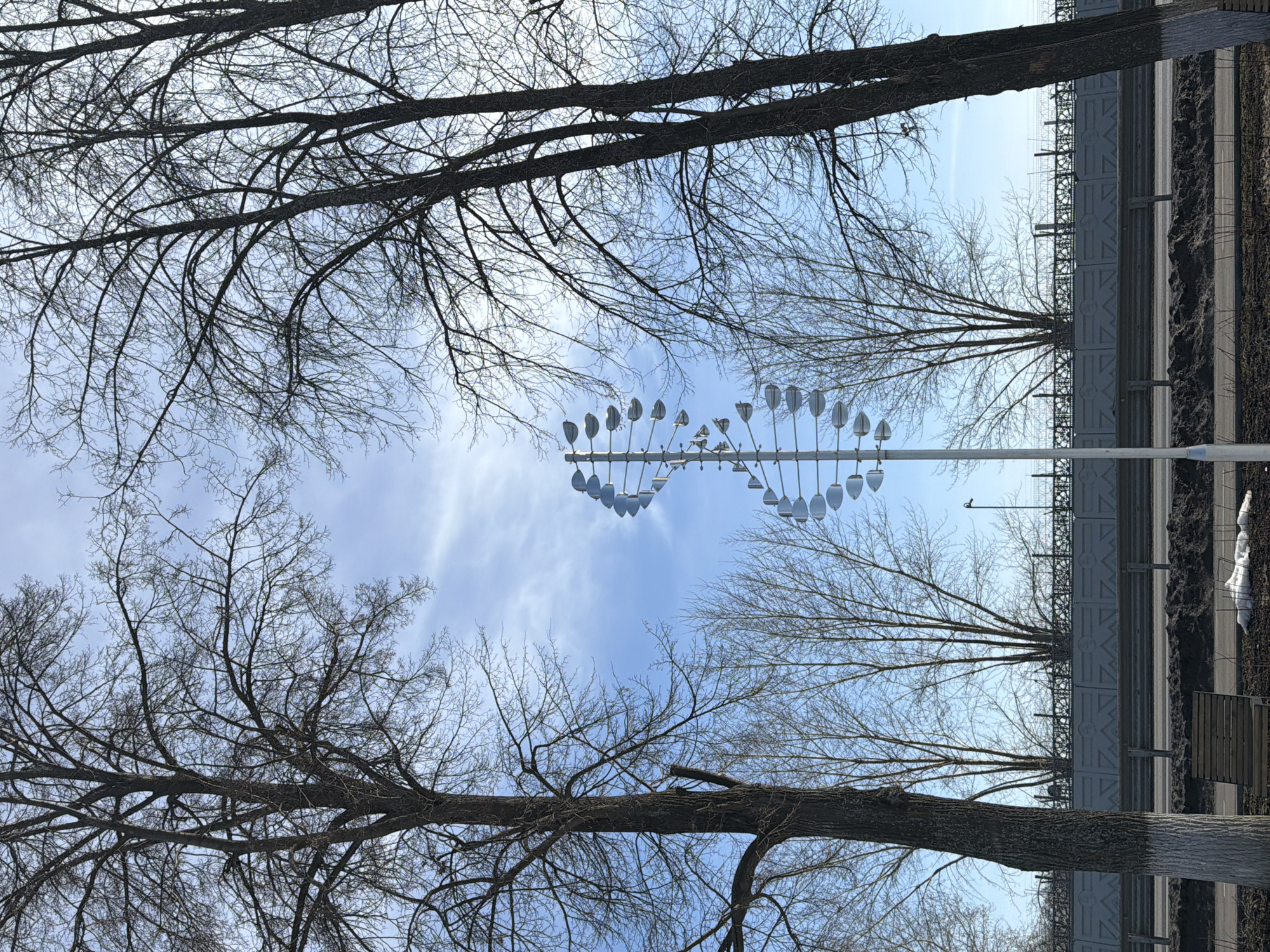 The image presents a view of a park or green space in Votkinsk, Russia, framed by bare tree branches silhouetted against a blue sky with wispy clouds. At the center of the image, there's a modern wind chime or kinetic sculpture consisting of multiple metallic, tulip-shaped elements suspended from a horizontal bar atop a white pole. In the background, a gray sound barrier wall runs parallel to a paved road or pathway. Adjacent to the road, there is a narrow strip of soil and dead vegetation. The image is composed in such a way that the tree branches act as a natural frame, drawing the eye towards the wind chime and the sky beyond. The leafless trees indicate a cold season, likely late autumn or winter.