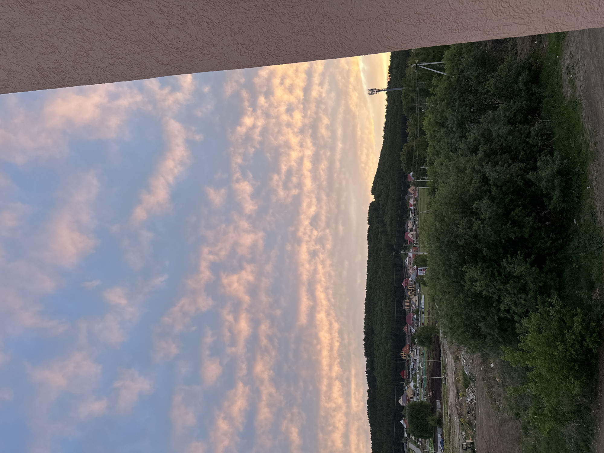 The image depicts a landscape with a blend of natural and built elements, partially framed by a building's edge. The sky is filled with light-colored, striated clouds, suggestive of early morning or late evening light. A dense line of trees forms a verdant barrier, with a few houses visible through the foliage along the hillside. A dirt or gravel path runs along the edge of the trees. A telecommunications tower is visible near the center of the horizon. The city of Unknown, Unknown is not recognizable in the image, but the houses hint at an urban or suburban setting.