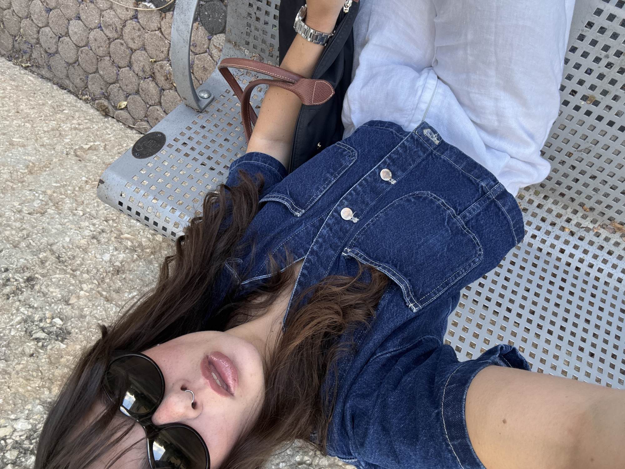 A woman with long, dark hair and sunglasses is sitting on a metal bench in Tel Aviv, Israel. She is wearing a blue denim jacket and white pants. A brown leather handbag is next to her, and she is wearing a silver watch on her left wrist. The bench is made of metal with a perforated pattern, and it appears to be in an outdoor setting with a textured stone ground.