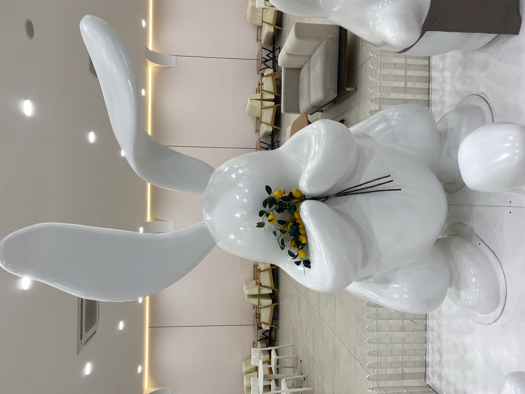 A large, white, abstract sculpture of a rabbit holding a bouquet of yellow lemons and green leaves stands prominently in the foreground. The rabbit has long, pointed ears and is positioned in front of a waiting area with light-colored sofas and chairs. In the background, there is a clean, modern interior with tiled floors and a pale pink wall. The scene suggests a public space like a cafe or a reception area. The image does not provide any information about the city of Da Nang, Vietnam.