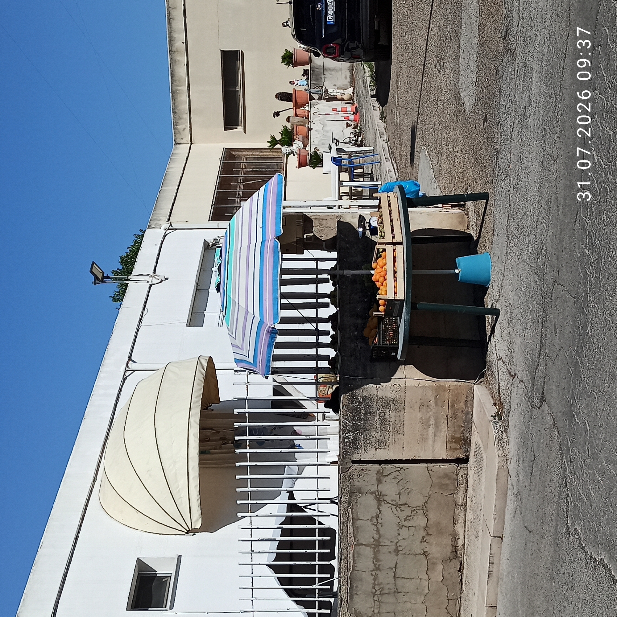 The image shows an outdoor fruit stand in Modugno, Italy, on a clear day. The stand features a table laden with crates of oranges and other fruits, partially covered by a colorful striped umbrella. A set of scales is visible on the right side of the table. Behind the fruit stand, a white building with a beige awning is visible, along with a white metal fence. Further back, potted plants and statues are arranged on a ledge. A black Fiat 500 car with the license plate GZ004ZV is parked to the right of the building. The ground is paved with asphalt. A timestamp in the bottom right corner reads "31.07.2026 09:37".