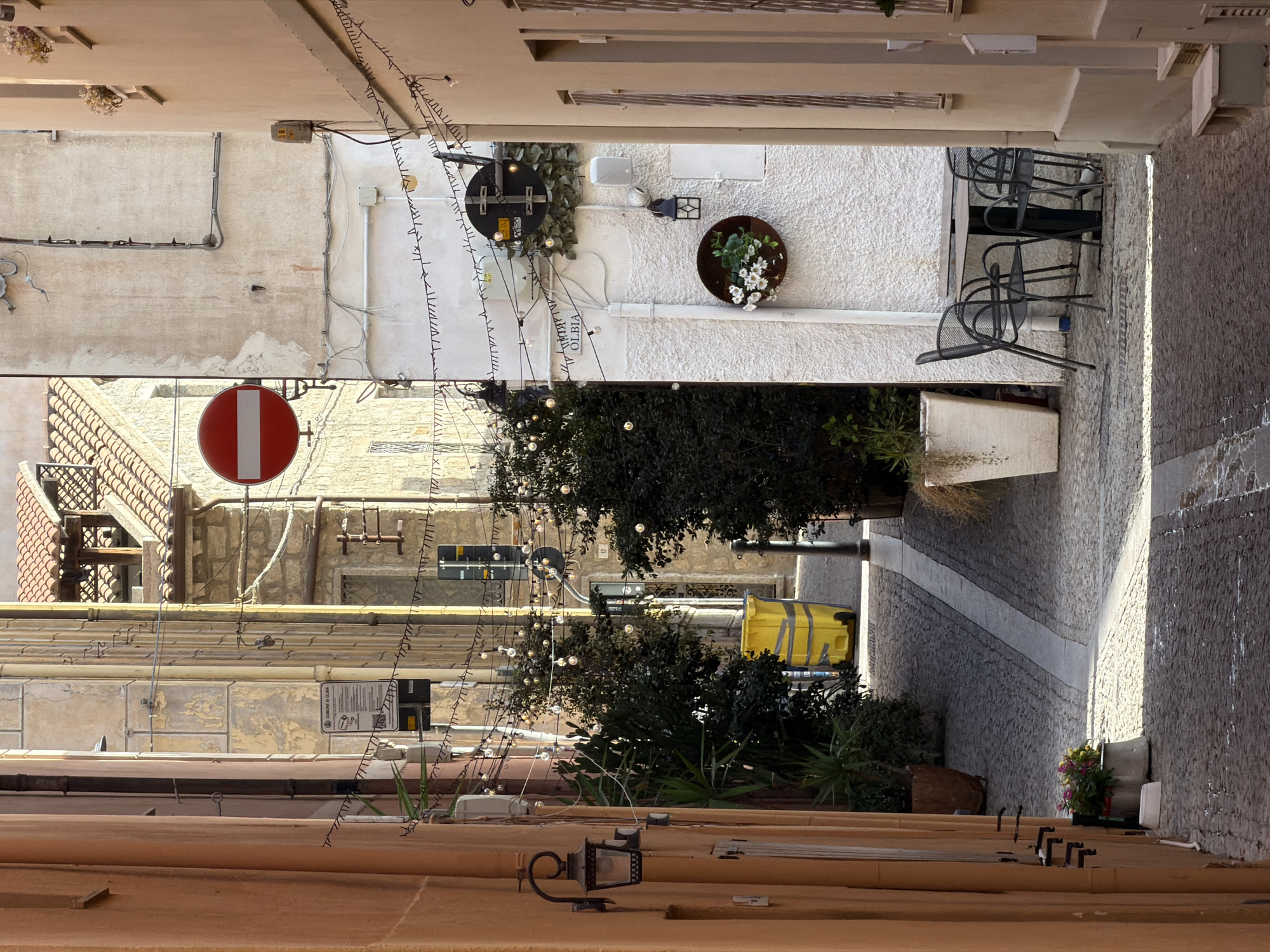 The image depicts a narrow, cobblestone alleyway in Olbia, Italy. On the left, an orange-colored building features a decorative lantern and a planter with flowers. The right side of the alley is dominated by a textured white building with an outdoor seating area consisting of a square table and four chairs. String lights are visible, draped across the alley above the seating area and extending further down the street. A circular planter with white flowers hangs on the white building. Further down the alley, a yellow trash receptacle is visible. Overhead, a red and white circular "no entry" traffic sign is mounted on a pole. Buildings with stone facades and red-tiled roofs are visible in the background.