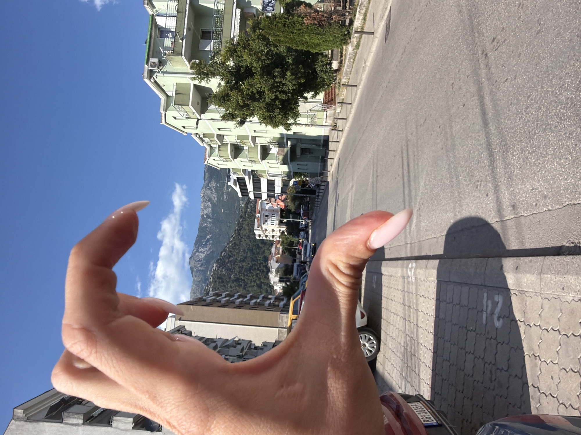 A first-person perspective shot shows a person's hand in the foreground, obscuring a view of a street scene. The hand, with long, light-pink acrylic nails, is positioned to frame the background. The background depicts a residential area with buildings of varying heights and architectural styles. A multi-story building with light green balconies dominates the left side of the frame. Further in the distance, a mountain range is visible under a clear blue sky with scattered white clouds. The street is paved with asphalt, and a sidewalk with interlocking gray pavers runs alongside it. Several cars are parked along the street, and some parking spots are marked with white numbers painted on the pavers. The sun is casting strong shadows, suggesting it is midday. The overall impression is a bright, sunny day in a coastal town.
The image is oriented vertically. The presence of buildings, a paved road, and parked cars indicates an urban or semi-urban environment. The distant mountains suggest a location with a dramatic natural backdrop. The clear sky and bright sunlight point to fair weather. The cropped hints provided include details such as the clear sky, the green building with balconies, the asphalt road, the sidewalk pavers, and parts of the person's hand and nails. One hint also shows a car with a license plate that appears to have a Cyrillic alphabet, which is consistent with the location of Budva, Montenegro.