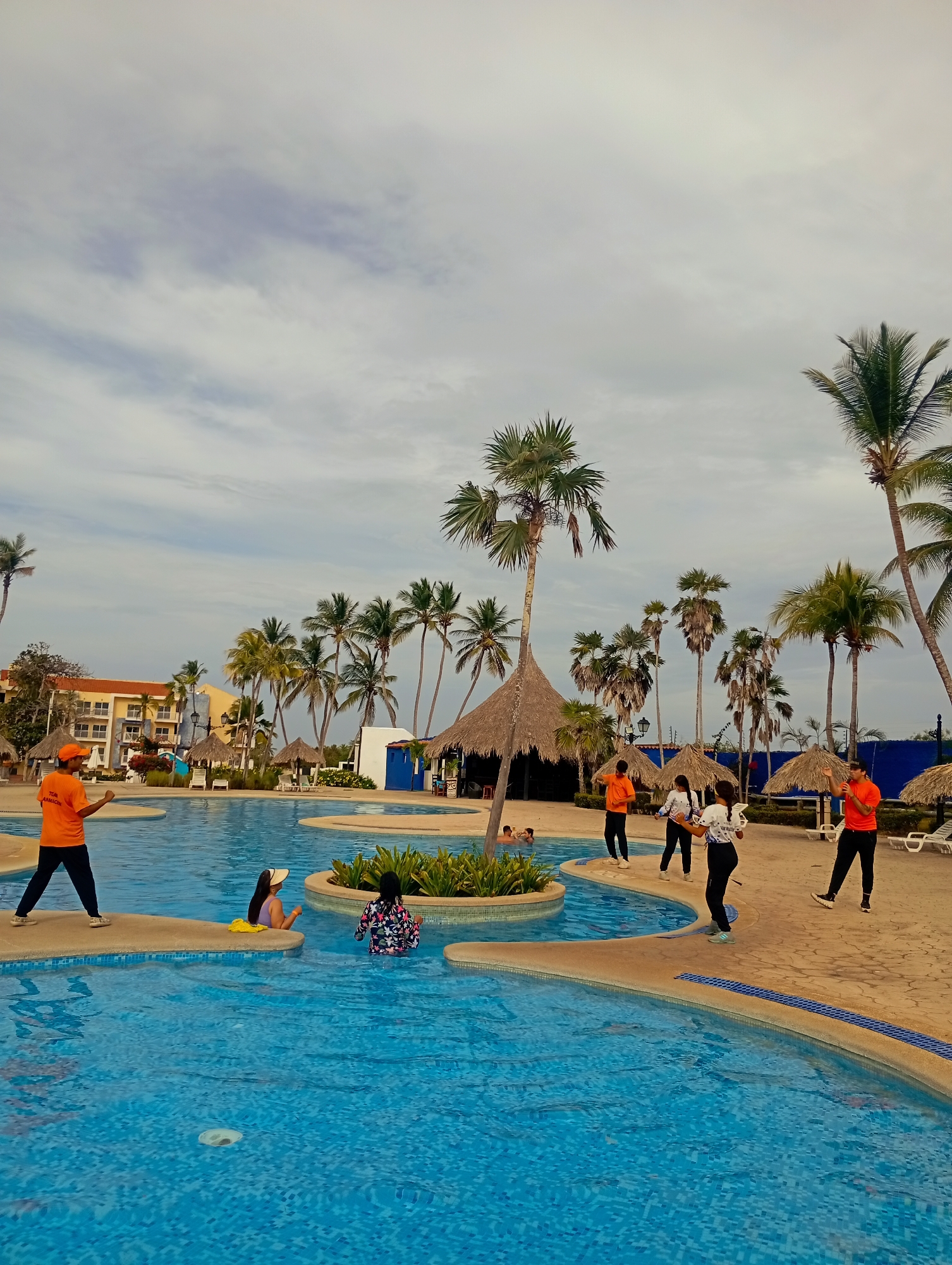 The image shows a resort pool area in Playa El Agua, Venezuela. A large, winding pool with blue tiled bottom is surrounded by palm trees and thatched-roof cabanas. Several people are in and around the pool, including two women sitting on a pool edge. Three staff members in orange shirts are standing on the poolside, seemingly interacting with a woman in a white t-shirt. The sky is overcast. A multi-story hotel building is visible in the background.