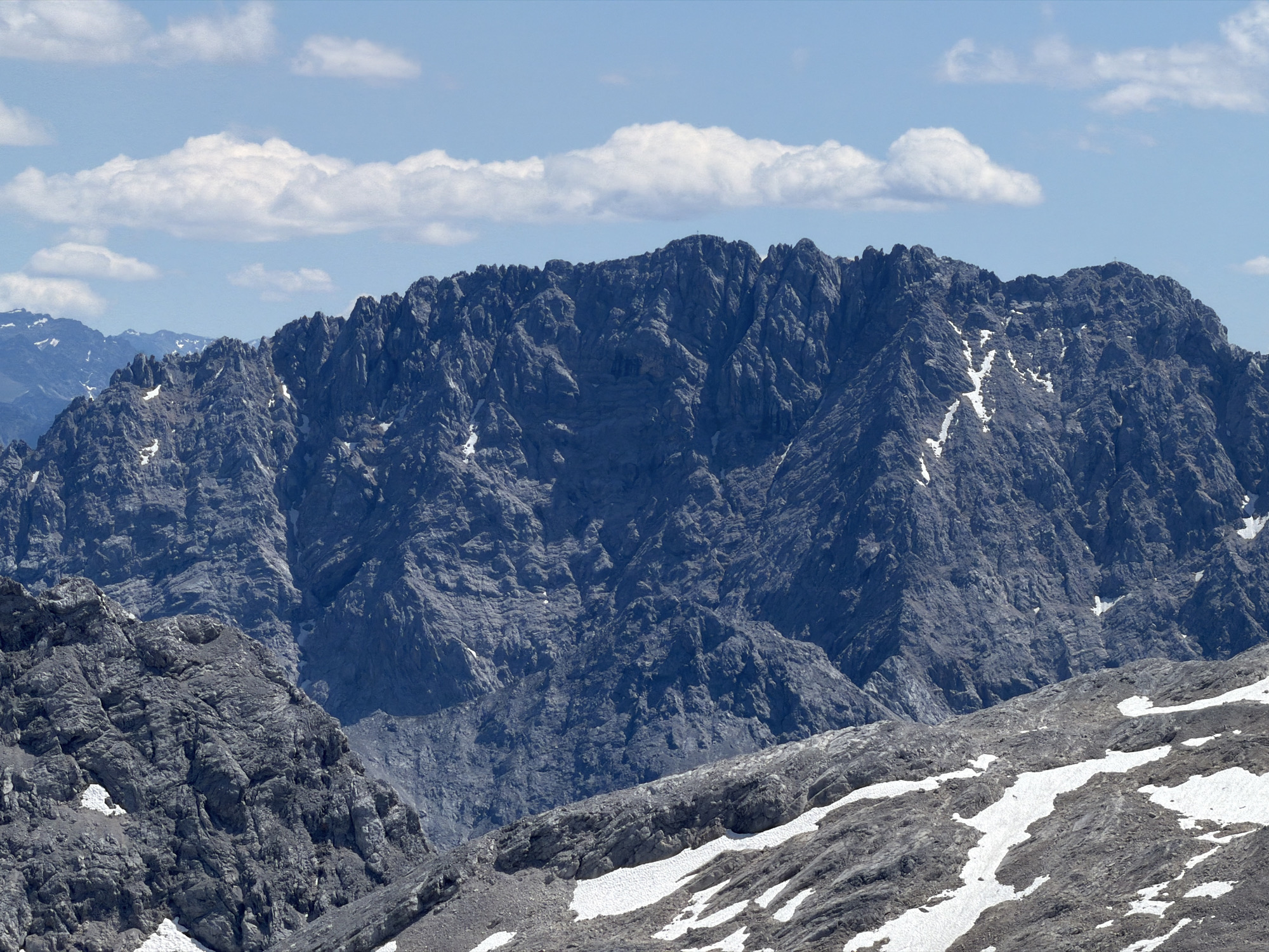 The image shows a panoramic view of the Zugspitze massif, the highest peak in Germany, located near Garmisch-Partenkirchen. The foreground is dominated by rocky slopes with patches of snow, indicating a high-altitude mountainous environment. In the middle ground, a jagged, dark gray mountain range stretches across the frame, its steep cliffs and textured surfaces clearly visible. The summit of this central mountain features a prominent cross. The background is a vast expanse of blue sky dotted with scattered white clouds. The overall scene is one of rugged, natural beauty, typical of the Bavarian Alps.
