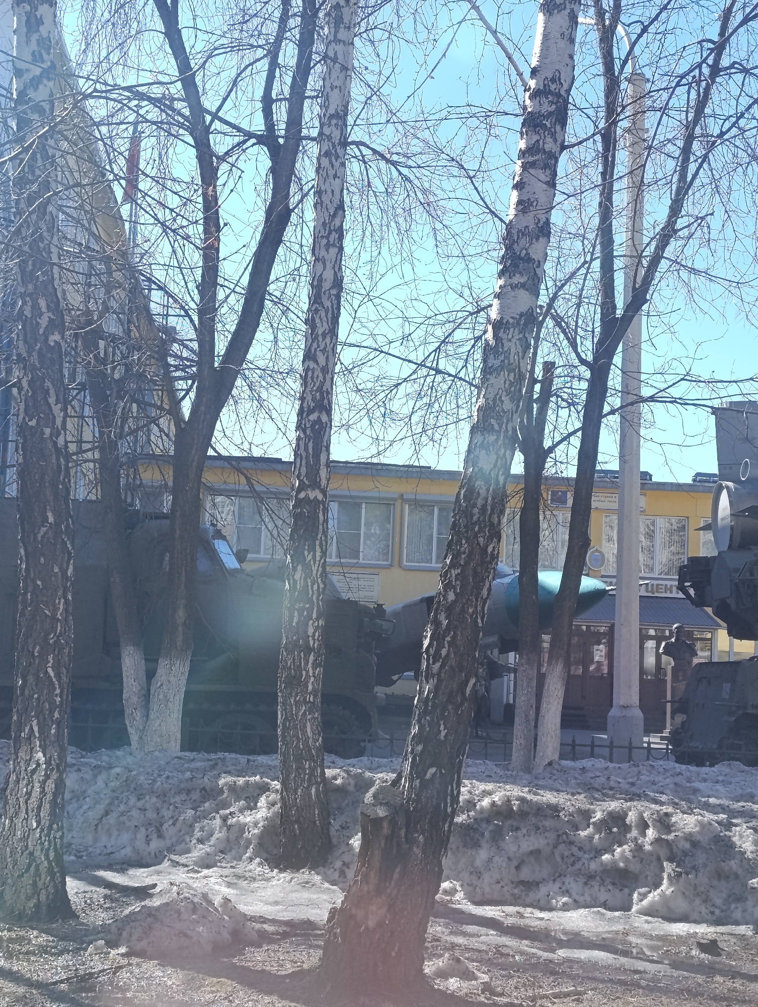 The image shows a snow-covered ground with several bare birch trees in the foreground. Behind the trees, there is a yellow two-story building with windows and a sign in Russian. Military equipment, including tanks and possibly a plane or missile, are partially visible among the trees and in front of the building. The sky is light blue, indicating a bright day. The image suggests a military memorial or museum. Based on the available details, this is likely Victory Park in Krasnoyarsk, Russia, showcasing military vehicles.