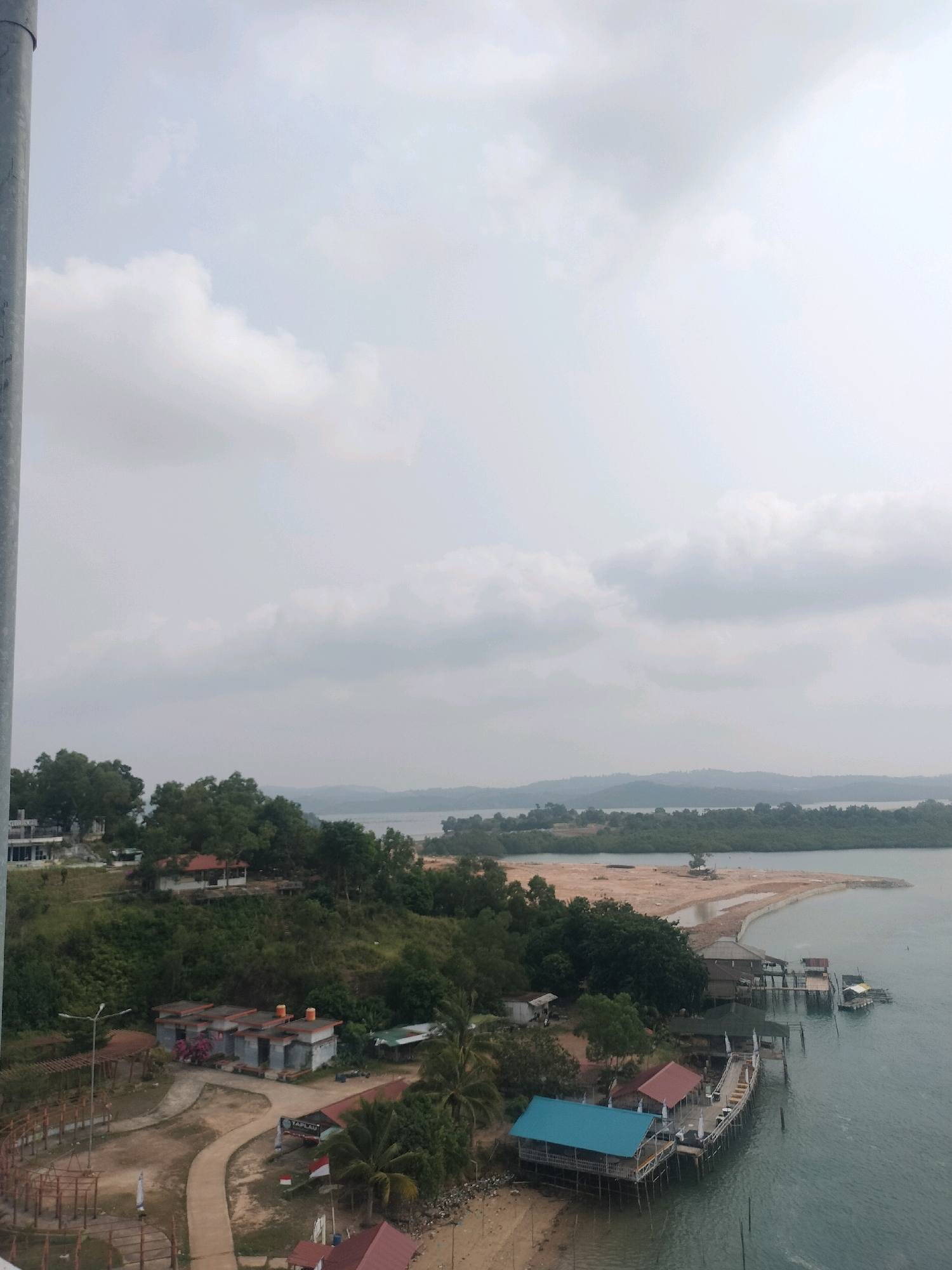 This aerial photograph captures a coastal landscape in Batam, Indonesia. In the foreground, a large metal pole is visible on the left. A paved pathway winds through a small settlement with several buildings. Some of these buildings appear to be small houses or bungalows with red and grey roofs. A white and red Indonesian flag is displayed prominently. Beyond the settlement, lush green trees and vegetation cover the hilly terrain.
The coastline features a sandy beach and several stilted structures, including jetties and waterfront buildings with colorful roofs, one with a distinctive blue roof. The water is a calm, greenish-blue. In the distance, a body of water stretches towards the horizon, with islands and hills dotting the landscape under a cloudy sky. The overall scene suggests a tropical, possibly tourist-oriented, coastal area with a mix of residential buildings, recreational facilities, and natural scenery.