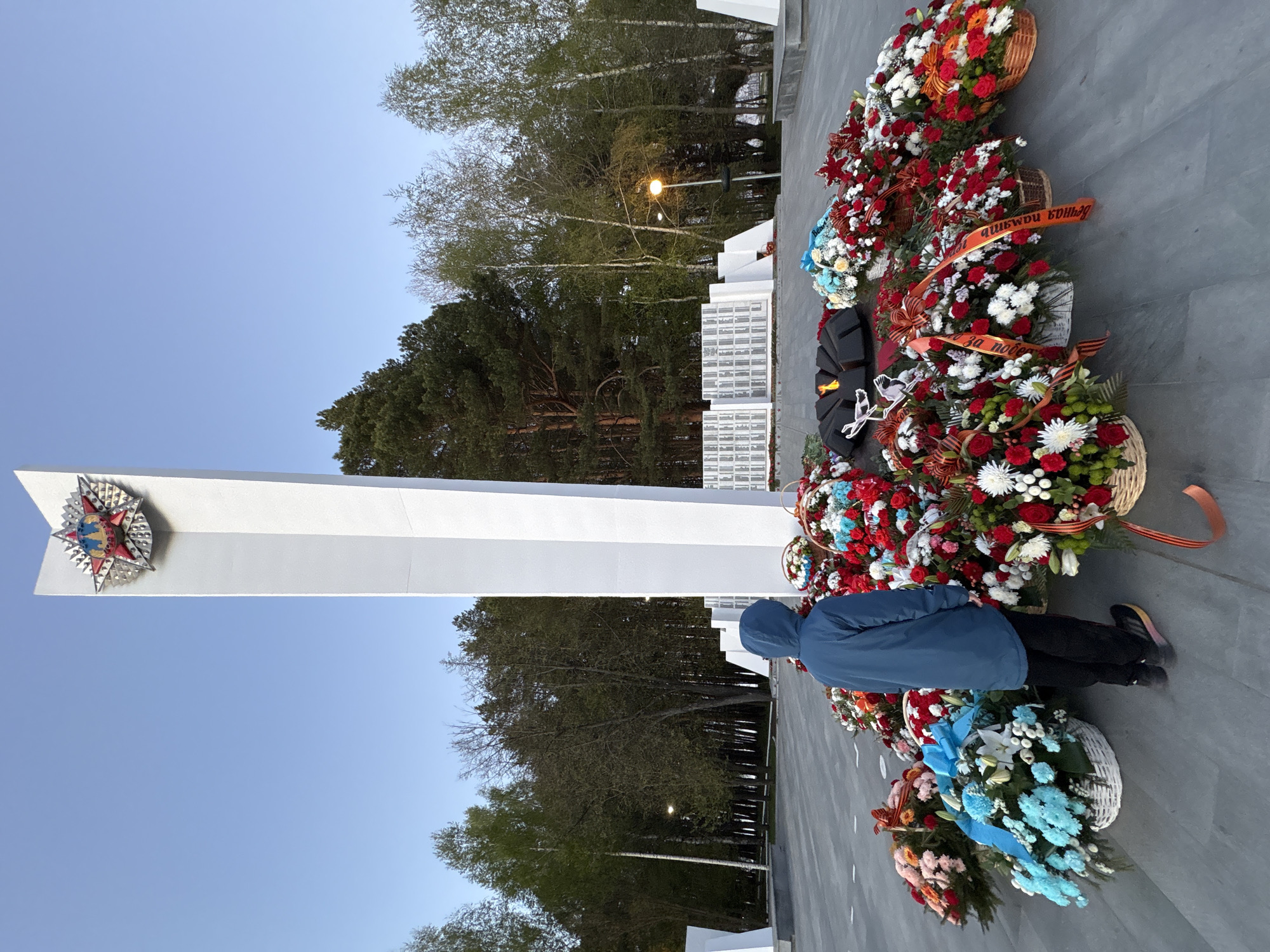 A tall white monument with a Soviet star on its face stands against a backdrop of trees and a clear, blue sky. The monument is located in Chaikovsky, Russia. A person wearing a blue hooded jacket is standing in front of the monument. Wreaths of flowers are arranged at the base of the monument.