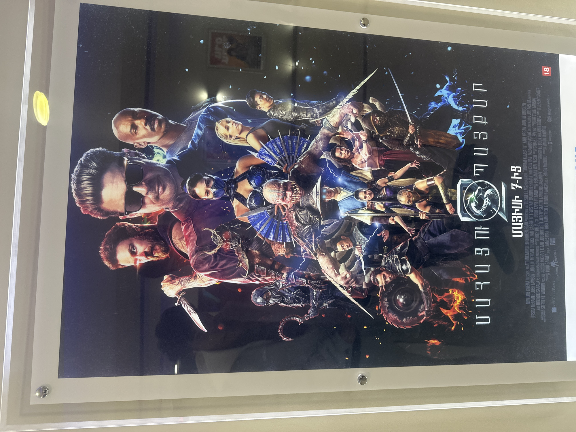 A movie poster for "Mortal Kombat" (2021) is displayed in a clear frame, captured at a 90-degree rotation clockwise. The poster features a dynamic collage of characters from the film set against a dark, cosmic-like background with subtle stars.
Prominently featured characters include Cole Young (center, wearing sunglasses), Sonya Blade (blonde, left), Jax (top left, bald), Kano (bottom left, bearded, holding a dagger), Mileena (bottom center, with blue fans), Sub-Zero (right of Mileena, emanating ice), Scorpion (top right, in samurai armor with a sword and fire effects), Liu Kang (bottom right, with fire effects), Raiden (right, with lightning effects), Kung Lao (top right, in a wide-brimmed hat), and Goro (bottom right, multi-armed with fire). The characters are arranged in action-oriented poses, wielding their signature weapons and powers, suggesting an intense battle or conflict. Their expressions are serious and determined.
Notable details include vibrant special effects such as ice, lightning, and fire surrounding the characters. The poster is encased in a reflective frame, indicating it's likely on display in an indoor public setting like a cinema.
Visible text on the poster, primarily in English and Armenian, includes:
- The main title "MORTAL KOMBAT" in English and "ՄՈՐԹԱԼ ԿՈՄԲԱՏ" in Armenian (written vertically).
- "ՄԱՅԻՍԻ 7-ԻՑ" (From May 7th) indicating a release date.
- Production companies: "NEW LINE CINEMA", "AN ATOMIC MONSTER/BROKEN ROAD PRODUCTION", "A FIRESIDE FILMS PRODUCTION".
- Cast credits for Jessica McNamee, Josh Lawson, Ludi Lin, Mehcad Brooks, Tati Gabrielle, Lewis Tan, Damon Herriman, Chin Han, Tadanobu Asano as Lord Raiden, Joe Taslim as Bi-Han, and Hiroyuki Sanada as Hanzo Hasashi and Scorpion. (Note: "KARL URBAN ADELINE RUDOLPH" also appears in the credit block, which is an unusual inclusion given the film's cast).
- Crew credits including Simon McQuoid (director), Stephen F. Windon (cinematography), and producers James Wan and Todd Garner.
- Logos for "18" (age rating), "WARNER BROS", "ATOMIC MONSTER", and "NEW LINE CINEMA".