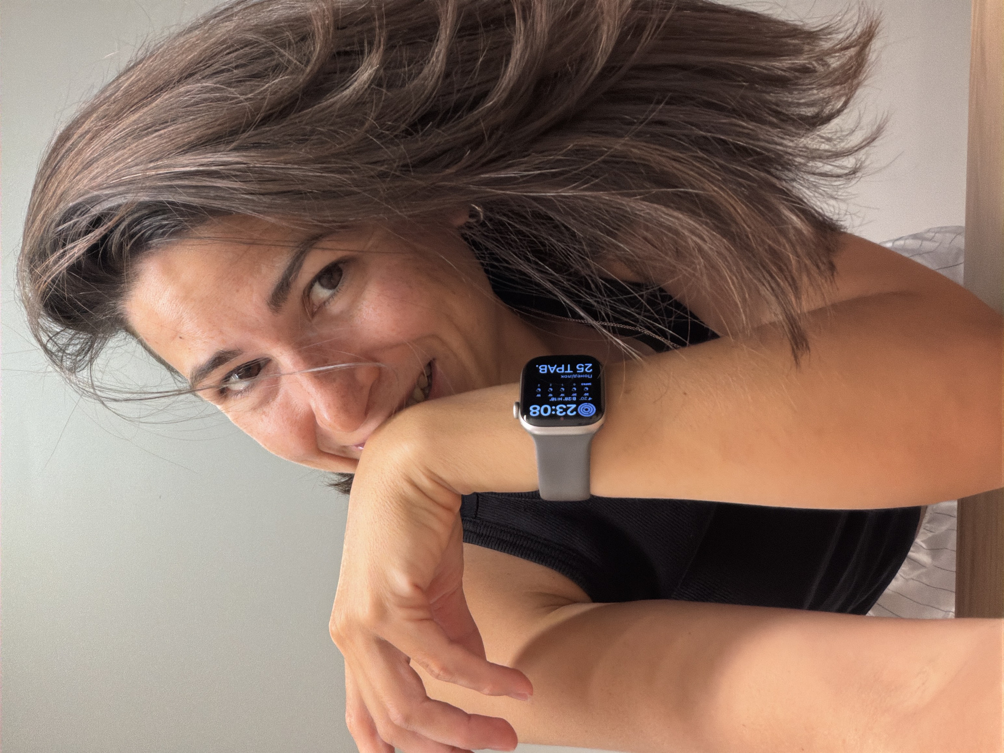 A young woman is holding her wrist up to the camera, showing off her Apple Watch. The watch face displays a digital time of 23:08 and the date "25 TPAV", indicating it might be set to a language other than English. The strap of the watch is grey. The woman is looking directly into the camera with a slight smile. Her hair is brown and casually styled. She is wearing a dark, possibly black, top. The background is a plain light color, likely a wall, and a glimpse of a striped fabric and a wooden surface are visible to the right. There are no obvious indicators of the location being Budva, Montenegro.