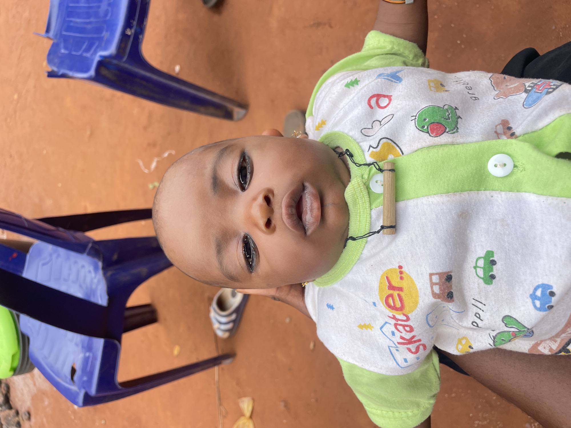 A baby is being held while lying on their back, angled towards the viewer. The baby is wearing a white and green patterned onesie. Two blue plastic chairs are visible in the background. The ground is an orange-brown color. The location is Ogbe, Nigeria.