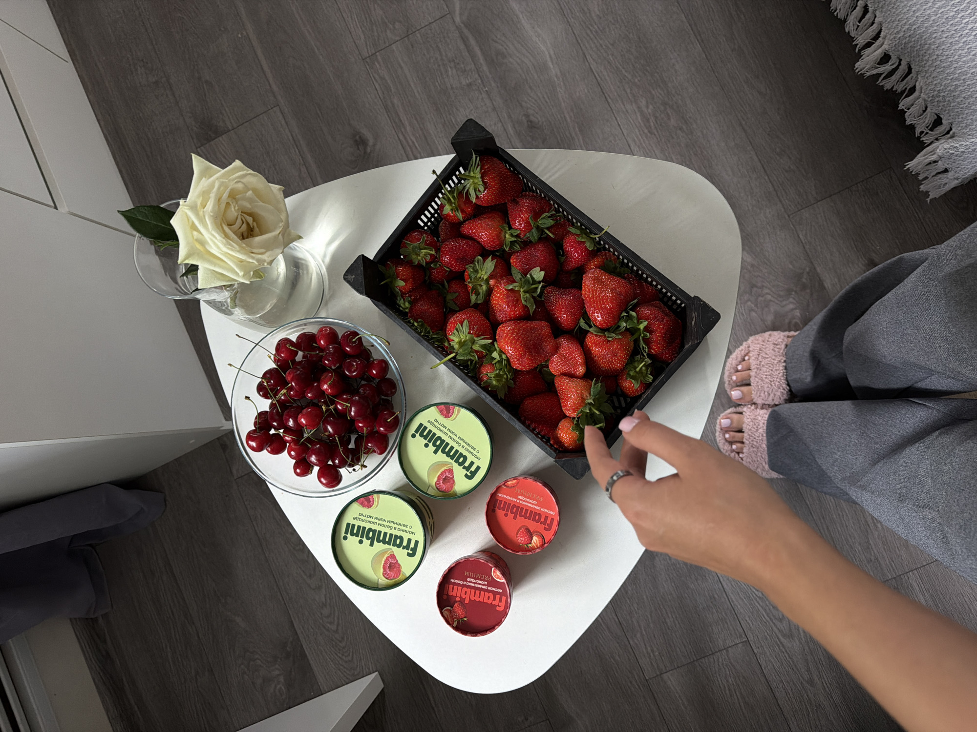 Overhead shot of a low white table situated on a dark wood floor, likely indoors. On the table are a vase with a single white rose, a glass bowl filled with cherries, a black crate brimming with strawberries, and four circular containers of Frambini, a chocolate brand. A woman's arm reaches into the frame, her hand with a ring pointing toward the strawberries. She is wearing grey pants and fuzzy pink slippers. The scene is suggestive of a casual gathering or personal indulgence with fresh fruit and chocolates. No specific details indicate the location as Kazan, Russia.