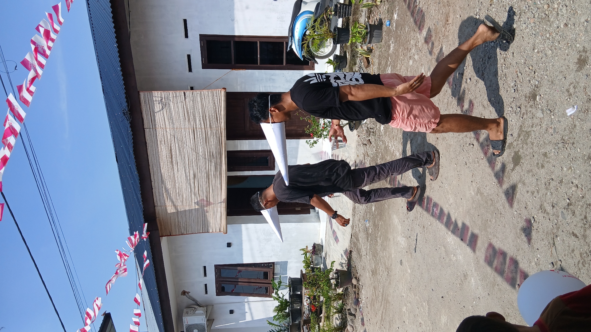 Two men walk along a dusty path in Klambir Lima Kebun, Indonesia. Both men are wearing large, white cone-shaped objects attached to their heads, obscuring their peripheral vision. The man on the left wears a black t-shirt and dark pants, while the man on the right wears a black t-shirt, pink shorts, and sunglasses. He is holding a clear plastic cup filled with a clear liquid. Behind them is a white building with a dark roof and a bamboo-covered window. Red and white striped banners are strung across power lines overhead. Potted plants are visible in front of the building and to the right of the path. A motorcycle is parked to the right of the building. The ground is a mix of dirt and gravel, with some red markings on the ground casting shadows. The sky is clear and blue.