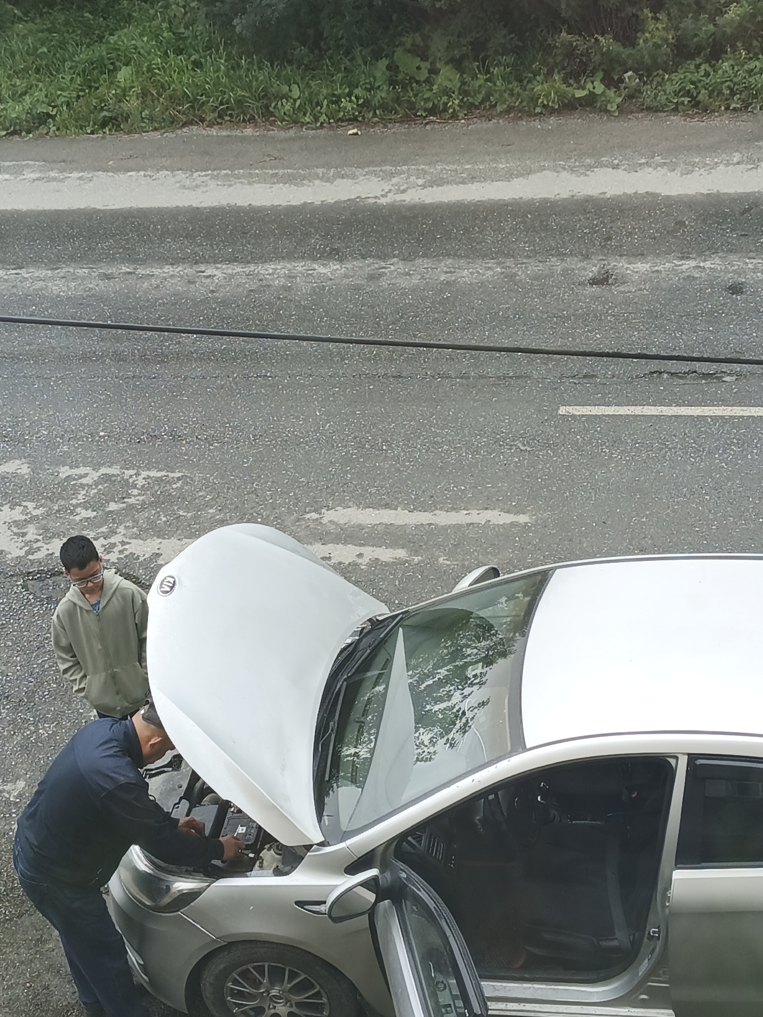 Two men are working on the engine of a silver car with its hood open. One man, wearing a dark blue long-sleeved shirt and jeans, is kneeling in front of the engine, appearing to be touching the battery. The other man, wearing a light olive green hooded jacket and glasses, stands beside the car, looking down at the engine. The car is parked on an asphalt road with white lane markings, bordered by a grassy embankment and trees. The scene appears to be a roadside location in the city of Zlatoust, Russia.