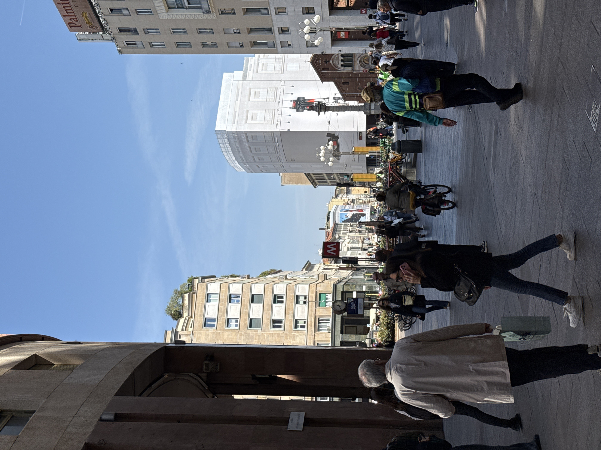 A ground-level shot presents a bustling city scene in Milan, Italy.
The street is populated with pedestrians of various ages and attire, walking in different directions. Some people are looking at their phones, while others are carrying bags.
Buildings flank both sides of the street, showcasing diverse architectural styles. Some are modern, while others appear historic. A large building is undergoing renovations with white protective coverings.
In the center of the scene, a red "M" sign is visible, indicative of the Milan Metro. Streetlights line the street, and bicycles are parked along the sidewalk.
The sky is clear and blue.
