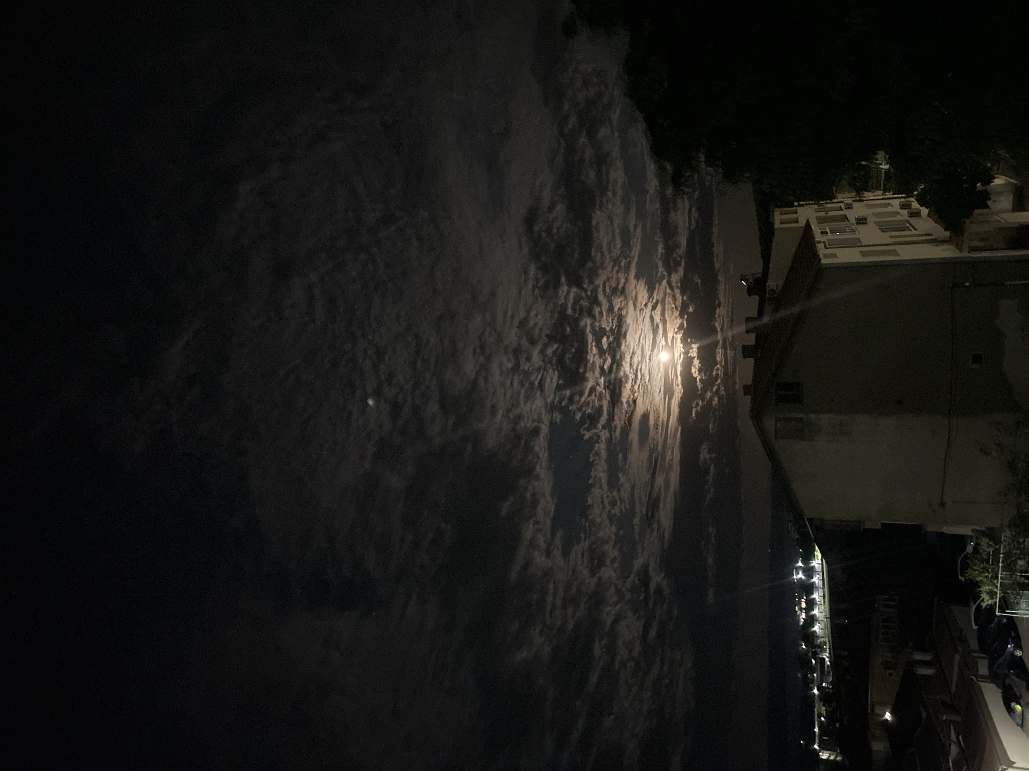 A nighttime scene, likely in an urban setting, depicts a view towards the sky and a building. The sky is dark, with a bright, possibly full moon, partially obscured by scattered clouds. Light from the moon reflects off the clouds, creating a hazy effect. To the right, a multi-story building is visible. The building appears to be made of stone or concrete and has a flat roof. Below the building, streetlights are visible along what appears to be a street. The overall scene is dark, and the lighting is low. The buildings and streetlights are near to a possible coast. The image may have been captured in the city of Burgas, Bulgaria.