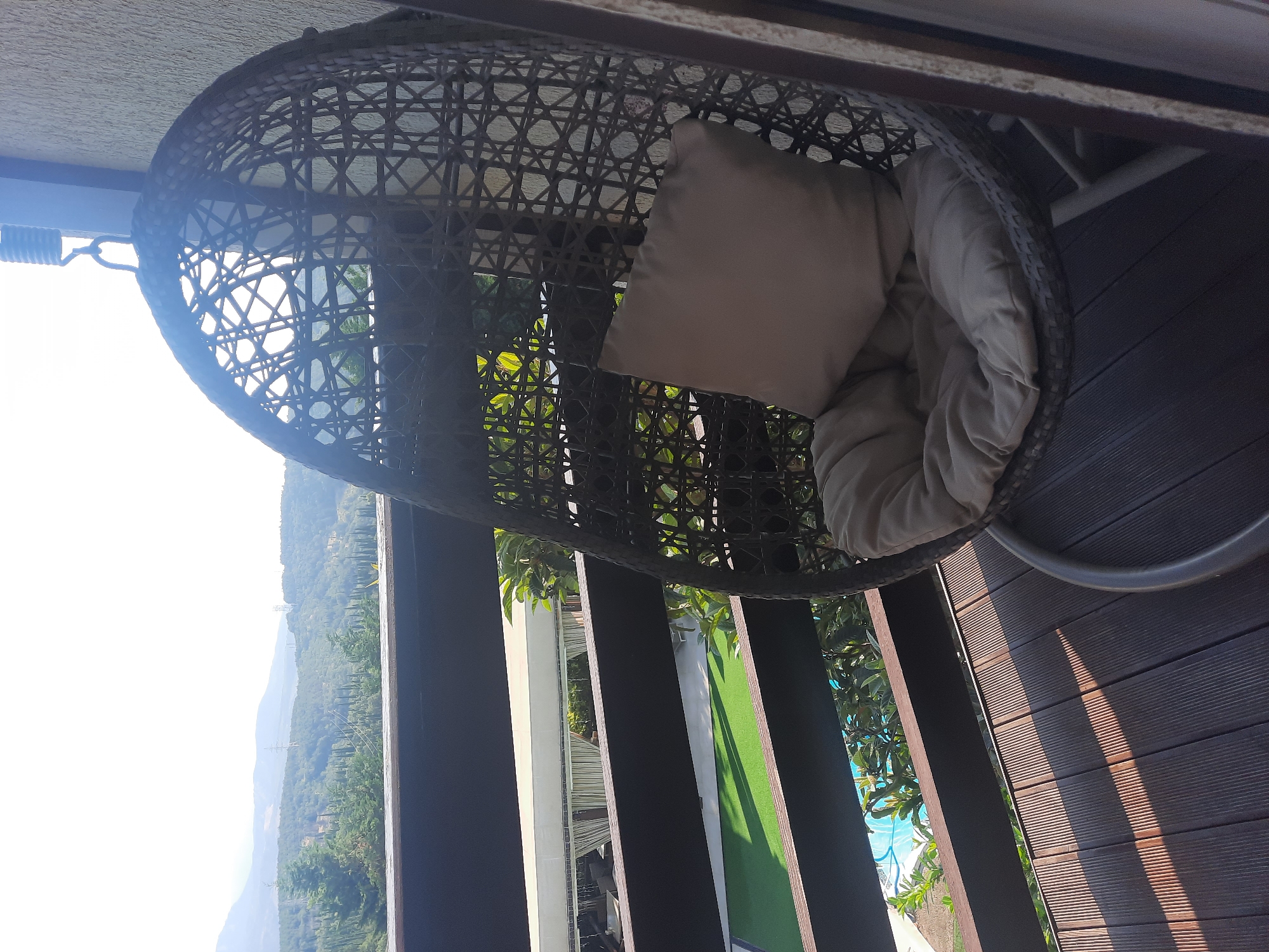 The image captures a close-up view of a hanging egg-shaped chair with a beige cushion and pillow, placed on a wooden deck. The chair is positioned on a balcony or patio that overlooks a lush, hilly landscape with trees and distant power lines under a bright, hazy sky. In the background, a swimming pool and some greenery are visible. The scene is illuminated by sunlight, casting shadows across the deck. No people are present in the image.