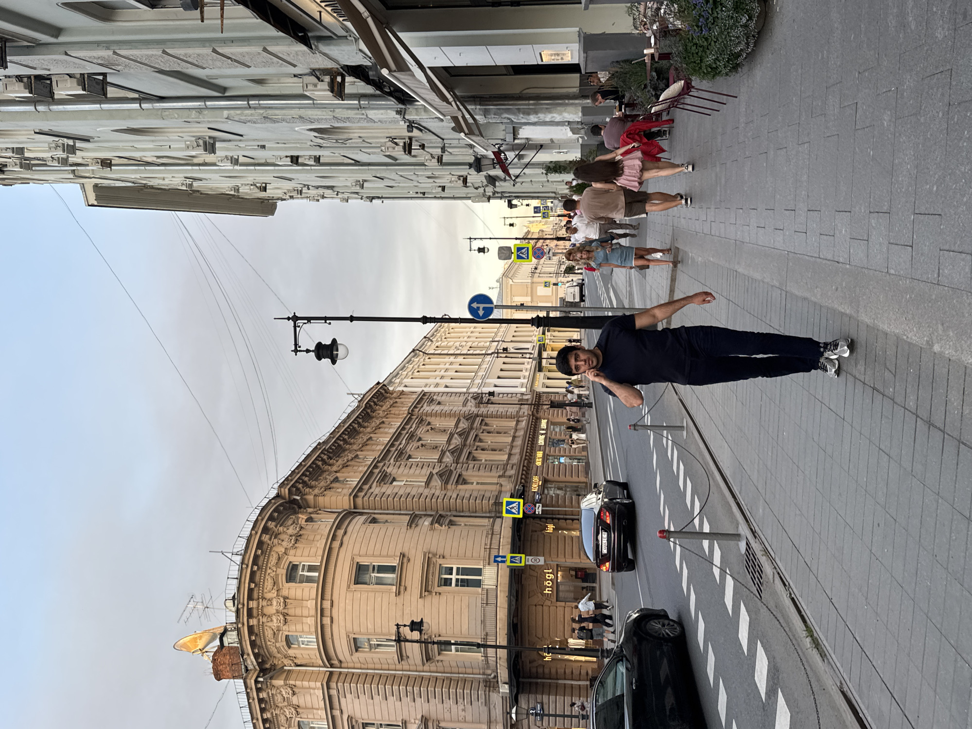The image depicts a street scene in Moscow, Russia, likely in the evening, given the lighting. The focus is on a man in the foreground walking on a sidewalk while talking on his phone. He is wearing a dark blue t-shirt and dark trousers, with grey sneakers. The sidewalk is paved with grey rectangular tiles.
To the left of the man, a black car is parked along the curb, and a dark sedan is driving away on the street, displaying a license plate with Russian characters and numbers. The street is marked with pedestrian crossings and metal bollards connected by chains.
The background features architectural elements characteristic of St. Petersburg, not Moscow, with ornate building facades. The building on the left is a corner building with intricate detailing and large windows, topped with a satellite dish. The building on the right is a pale, multi-story structure with many windows and air conditioning units, and a section of an awning suggests a commercial establishment, possibly a cafe or restaurant, with outdoor seating visible. Several other pedestrians are walking on the sidewalk in the distance. The sky is overcast with a soft, diffused light.