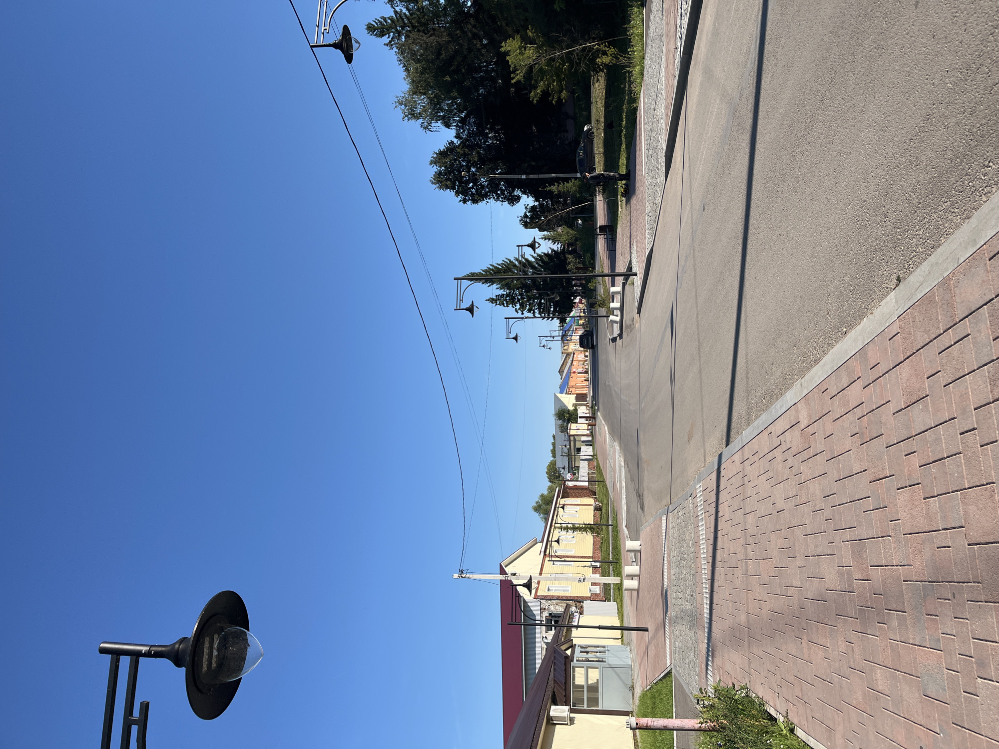 The image depicts a street scene in Blagoveshchensk, Russia, on a clear, sunny day. A paved road runs through the center of the frame, lined with a red brick sidewalk on the left and a stone-paved area transitioning to grass and trees on the right. Several lampposts with classic designs are strategically placed along the sidewalk. Buildings with light-colored facades and red roofs line the left side of the street. On the right, dense trees create a visual barrier. The sky is a vibrant, cloudless blue. There are no people or animals visible in the immediate scene. The overall impression is one of a quiet, well-maintained street in a town or small city.
