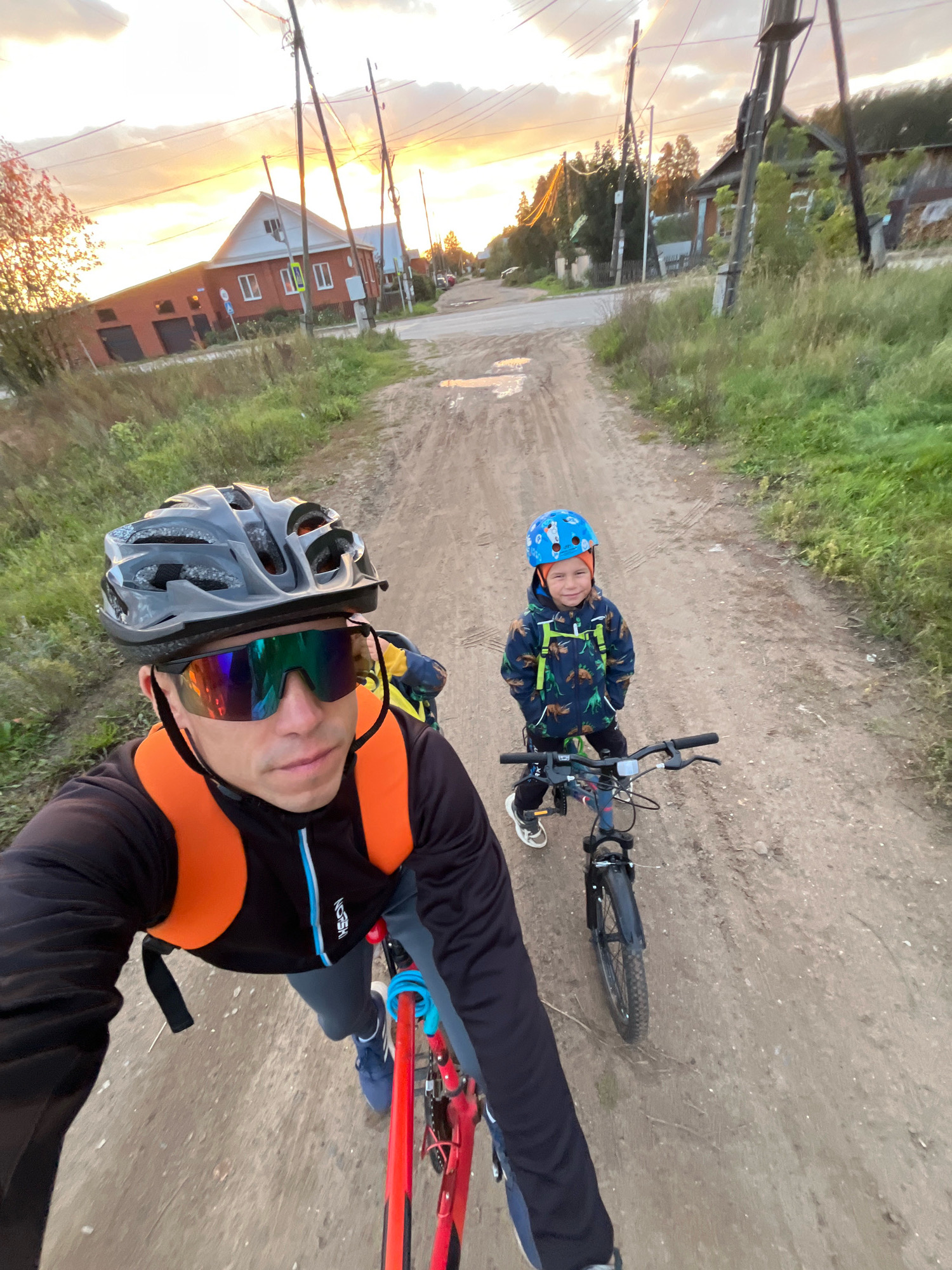 A high-angle, selfie-style shot captures a man and a child on bicycles on a dirt road at dusk in Chaikovsky, Russia. The man, wearing a grey helmet, reflective sunglasses, and an orange backpack over a black and grey cycling jacket, is in the foreground, facing forward. Beside him, slightly ahead and to his right, is a young boy wearing a blue helmet with a white pattern and an orange beanie underneath. The boy is dressed in a dark jacket with a dinosaur pattern and black pants, and is also on a bicycle.
The road is unpaved and dusty, with some patches of water or mud. Tall grass and weeds grow along the sides of the road. In the background, a rural landscape is visible with houses, power lines, and trees. The sky is a mixture of warm orange and yellow hues, indicating sunset. The overall atmosphere suggests a casual bike ride in a semi-rural or suburban setting.