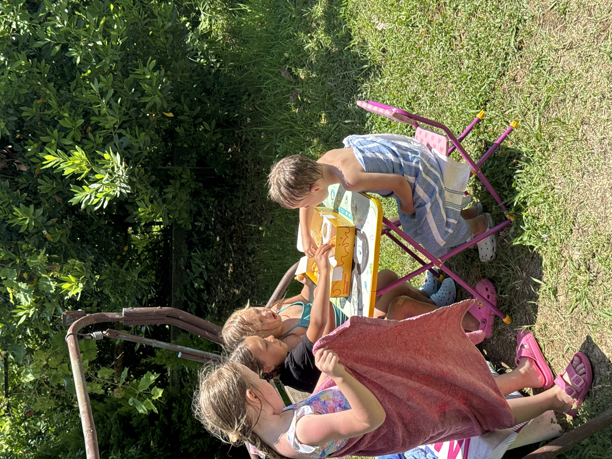 Four young girls are sitting at a small yellow table outdoors. One girl, on the far left, is standing and holding a pink towel. She is wearing a swimsuit and pink sandals. The second girl from the left is wearing a black t-shirt and is reaching for food from a yellow box on the table. The third girl from the left, in a teal swimsuit, is also reaching for food. The fourth girl, on the far right, is seated in a pink chair and is wrapped in a blue and white striped towel. She is looking at the food on the table. The table has a few items on it, including a yellow box, a white cup, and some food. The background consists of green grass and lush trees, suggesting a park or garden setting. The scene appears to be a sunny day. The city of Sochi, Russia, is known for its numerous parks and outdoor recreational areas, which would be consistent with this environment.