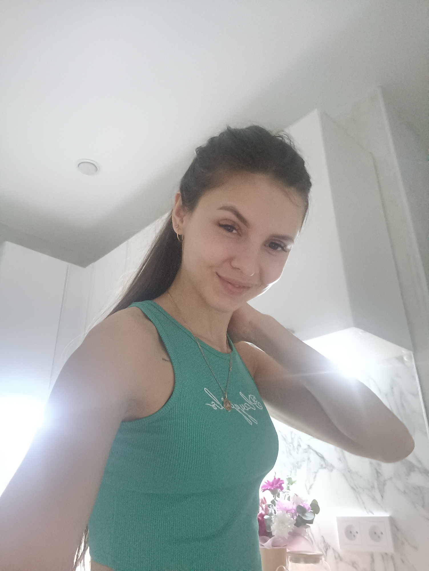 A selfie taken by a young woman with long brown hair, wearing a green tank top and a gold necklace, in an indoor setting. The background includes white cabinets, a marble wall, electrical outlets, and a vase of flowers on a counter. The location is Unknown, Unknown.