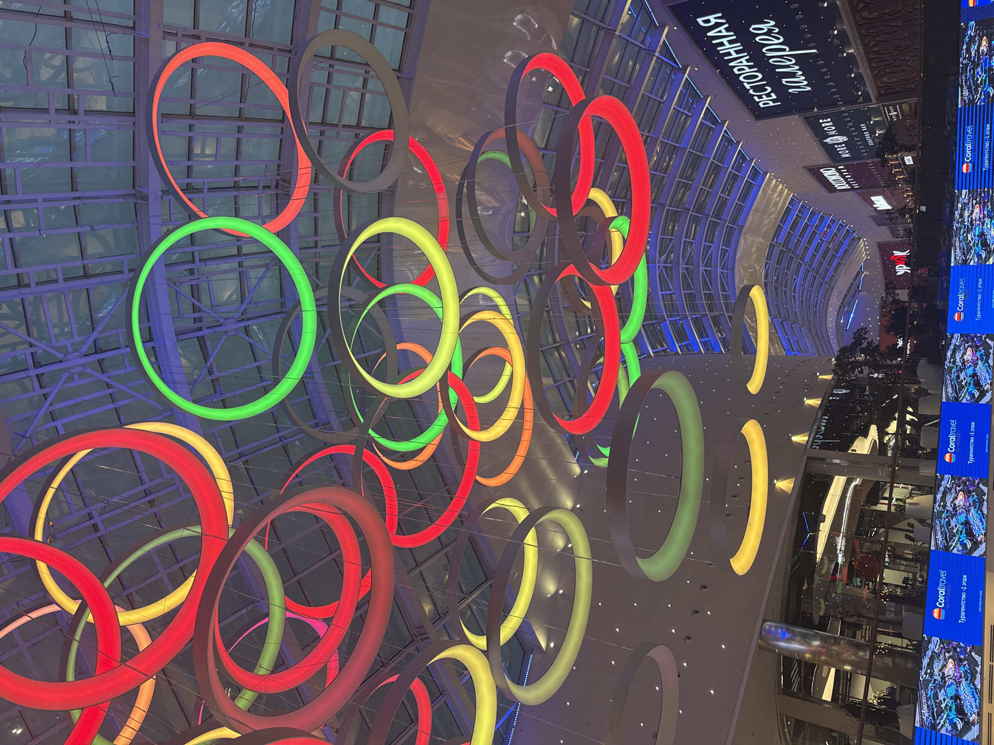 The image shows a brightly lit interior of a shopping mall, possibly in Khimki, Russia. Numerous circular, colored lights are suspended from the ceiling, creating a vibrant and modern ambiance. The lights are in various colors, including red, yellow, green, and pink, and are arranged in overlapping rings and clusters. The ceiling itself appears to be a glass or translucent material, allowing some natural light to filter through, and it is supported by a network of metal beams.
Below the lights, the mall's interior is visible, featuring shops and restaurants. Several signs are present, including one that reads "РЕСТОРАННАЯ галерея" (Restaurant Gallery) in large, illuminated letters. Other signs include "CORAL TRAVEL" and logos for various businesses. Large screens displaying images and text are positioned along the lower part of the image, featuring "Coral Travel" and Russian text.
The scene is generally clean and well-maintained, indicative of a commercial establishment. There are no visible people in the immediate foreground or clearly identifiable in the background. The overall impression is of a bustling and contemporary retail environment.