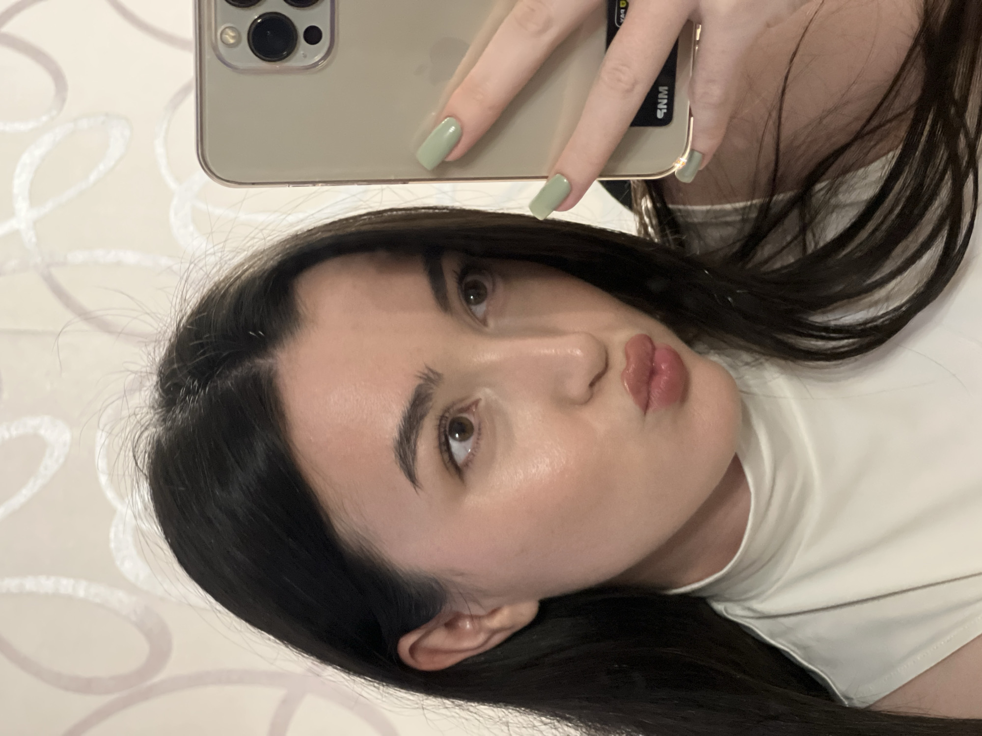 A young woman with long, dark hair takes a selfie in a mirror. She is wearing a white, sleeveless top with a high neckline. Her lips are pursed, and she is looking at the camera. Her fingernails are painted a light green color. In her right hand, she is holding a gold-colored smartphone, which is partially visible in the frame, showing a portion of the camera array. The background appears to be a wall with a subtle, light-colored wallpaper featuring a swirling, abstract pattern. The lighting is soft, casting a gentle glow on her face. No specific location in Kazan, Russia is identifiable from the image.