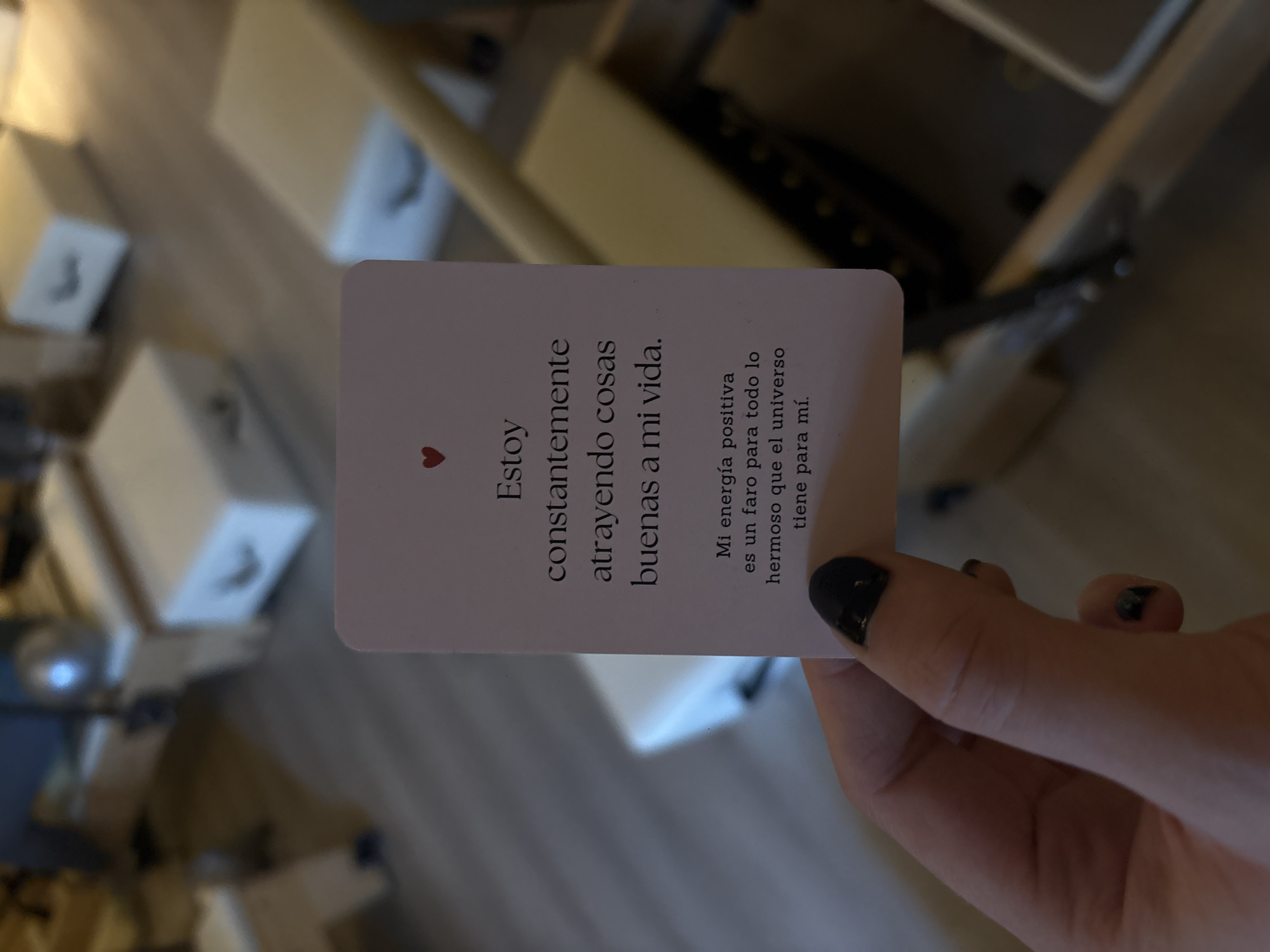 A hand holding a card with a Spanish affirmation. The card has a red heart and the text "Estoy constantemente atrayendo cosas buenas a mi vida. Mi energía positiva es un faro para todo lo hermoso que el universo tiene para mí." The hand is holding the card against a blurred background. The location is Mexico City, Mexico.