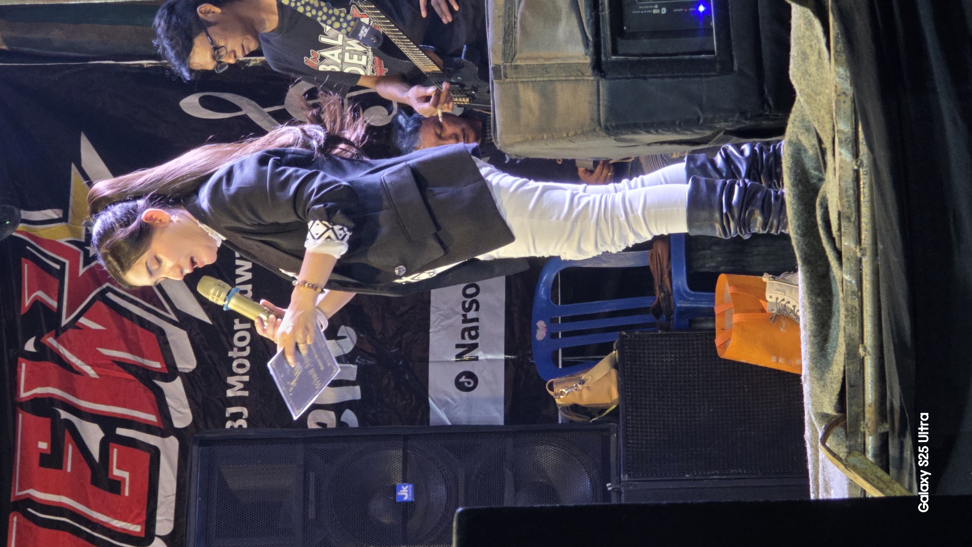 In Juwana, Indonesia, on a stage illuminated by spotlights, a woman stands center-frame, speaking into a golden microphone. She is dressed in a black blazer, white pants, and dark, ruched boots. She holds a small, rectangular plaque in her left hand, gesturing with it as she speaks. Behind her, a banner with bold red lettering is visible, partially obscured by a large black speaker to her left. To her right, a man wearing glasses and a black t-shirt, holding a guitar, looks towards the woman. Another person, partially visible behind the man with the guitar, is also on stage. In the foreground, an orange bag and a pair of beige boots rest near a blue chair. The overall scene suggests an outdoor event or performance.